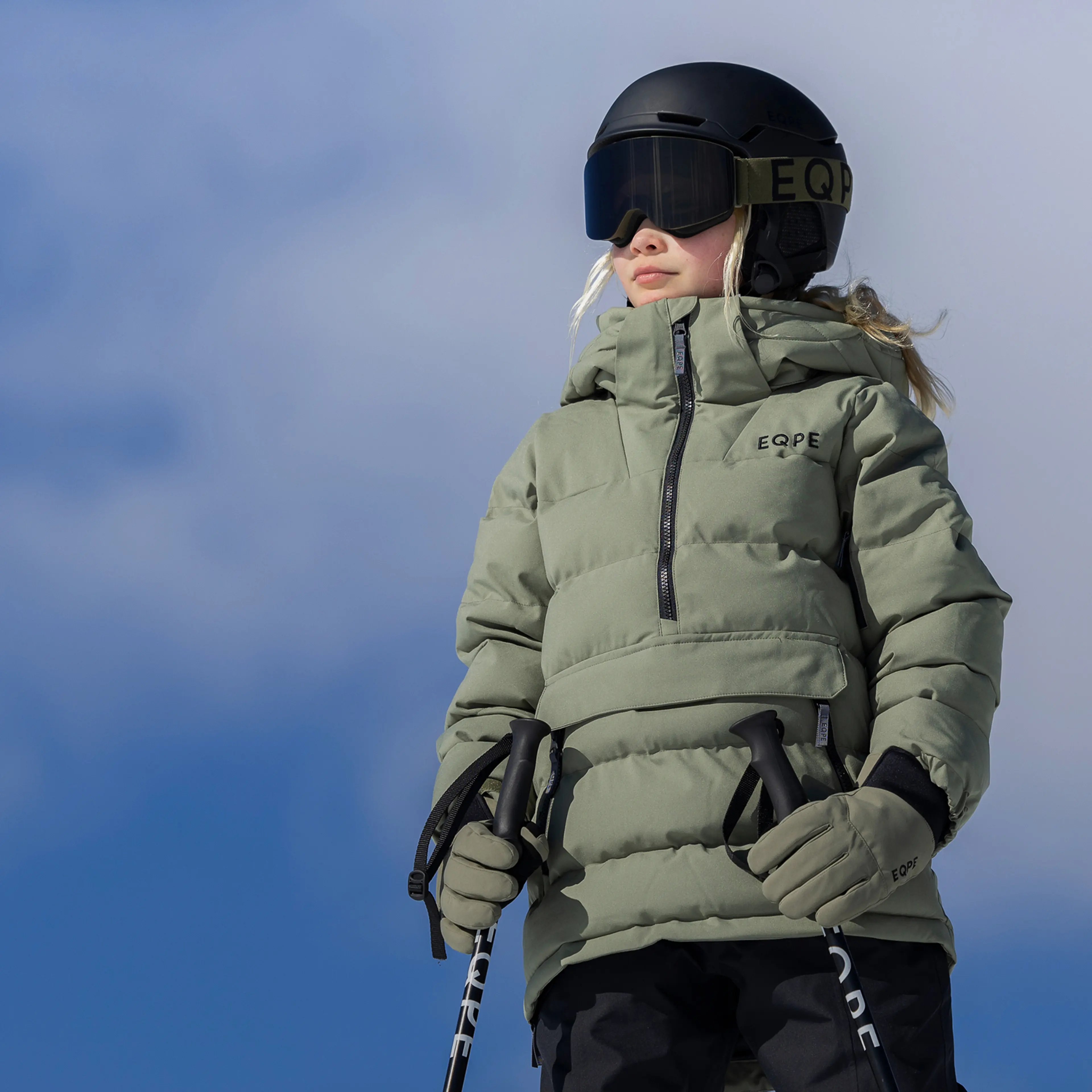 Child wearing a green puffer jacket and helmet, holding ski poles, stands against a clear blue sky.