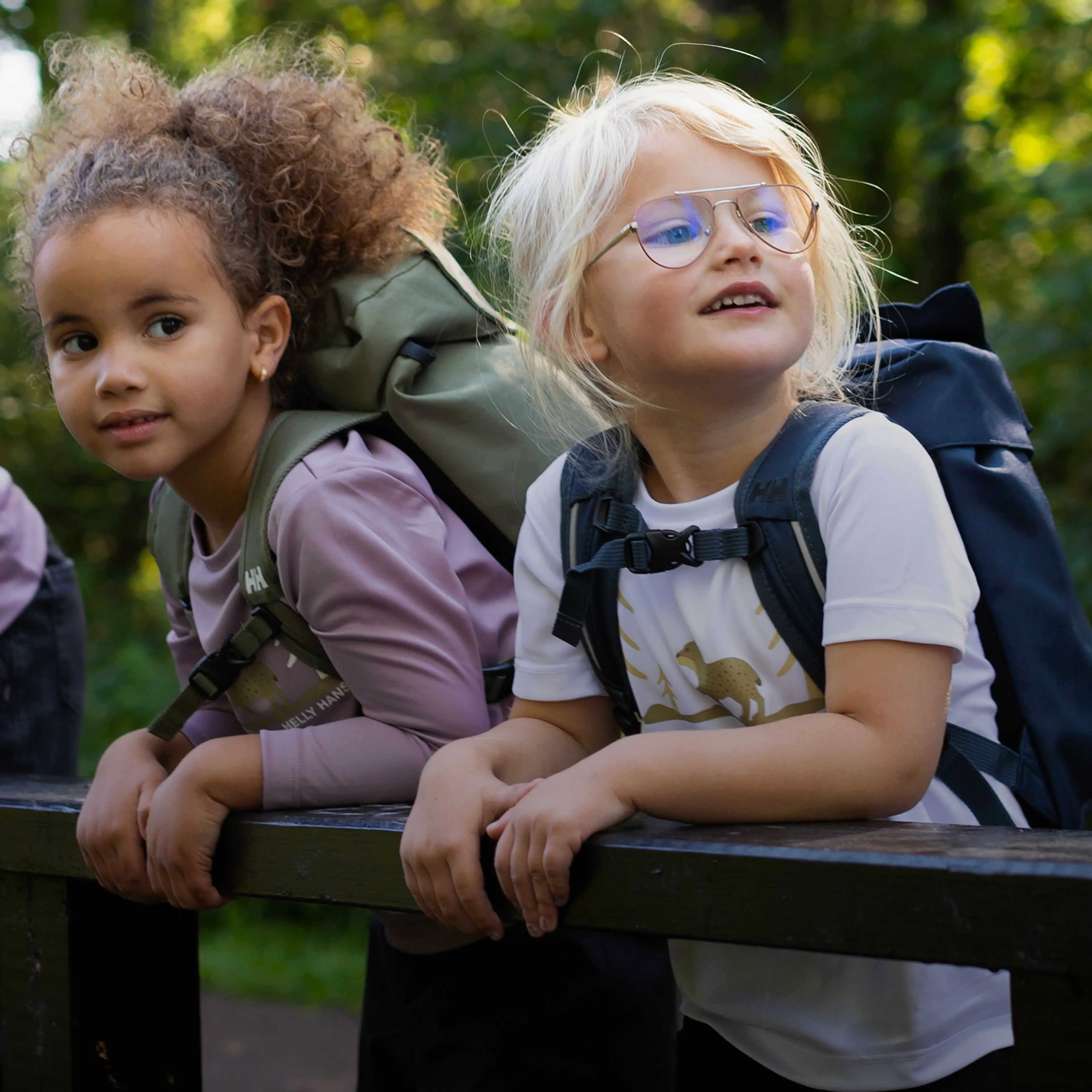 Two children with backpacks lean on a wooden railing, looking off into the distance in a green outdoor setting.
