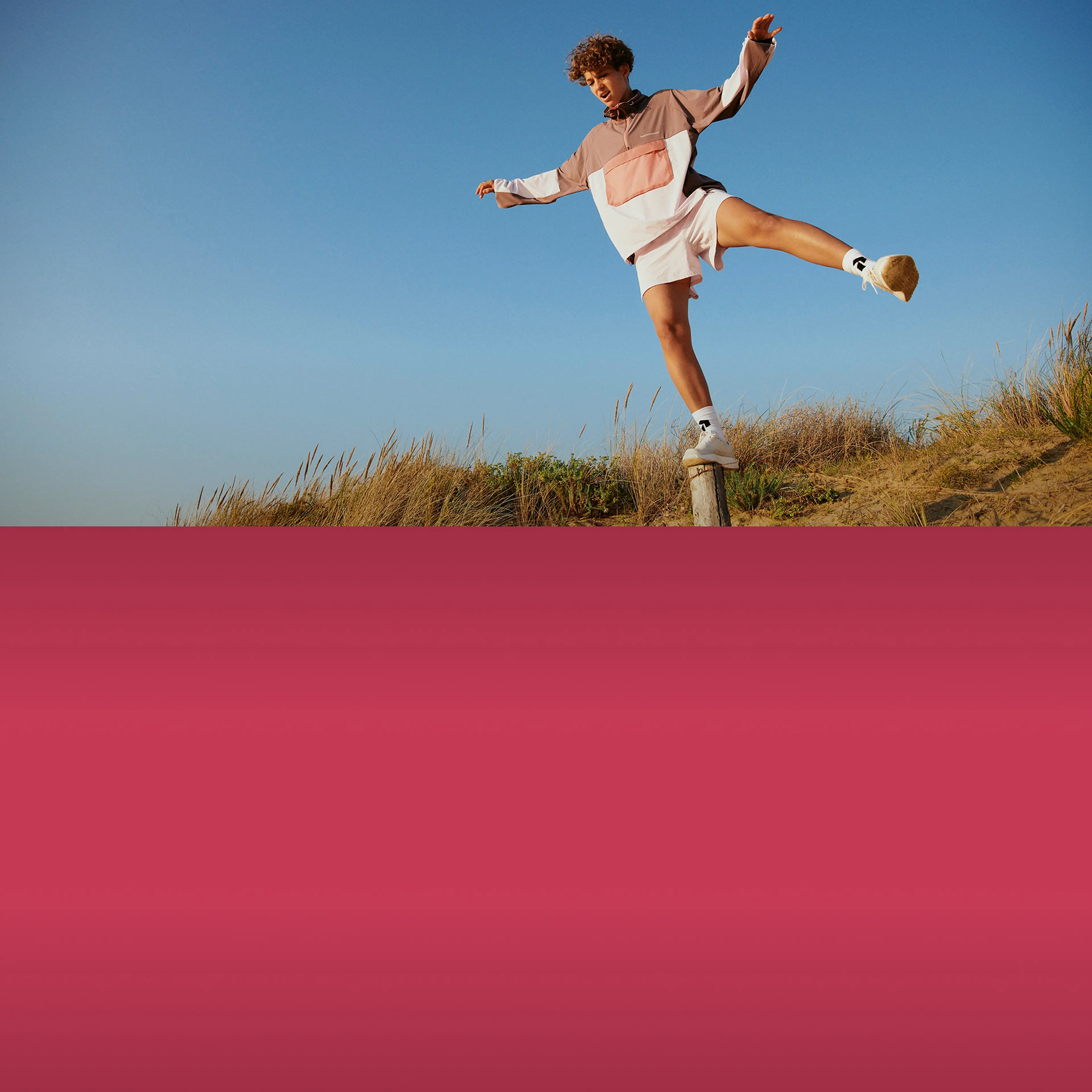 Person balancing on a wooden post in a grassy field, wearing a casual outfit, under a clear blue sky.