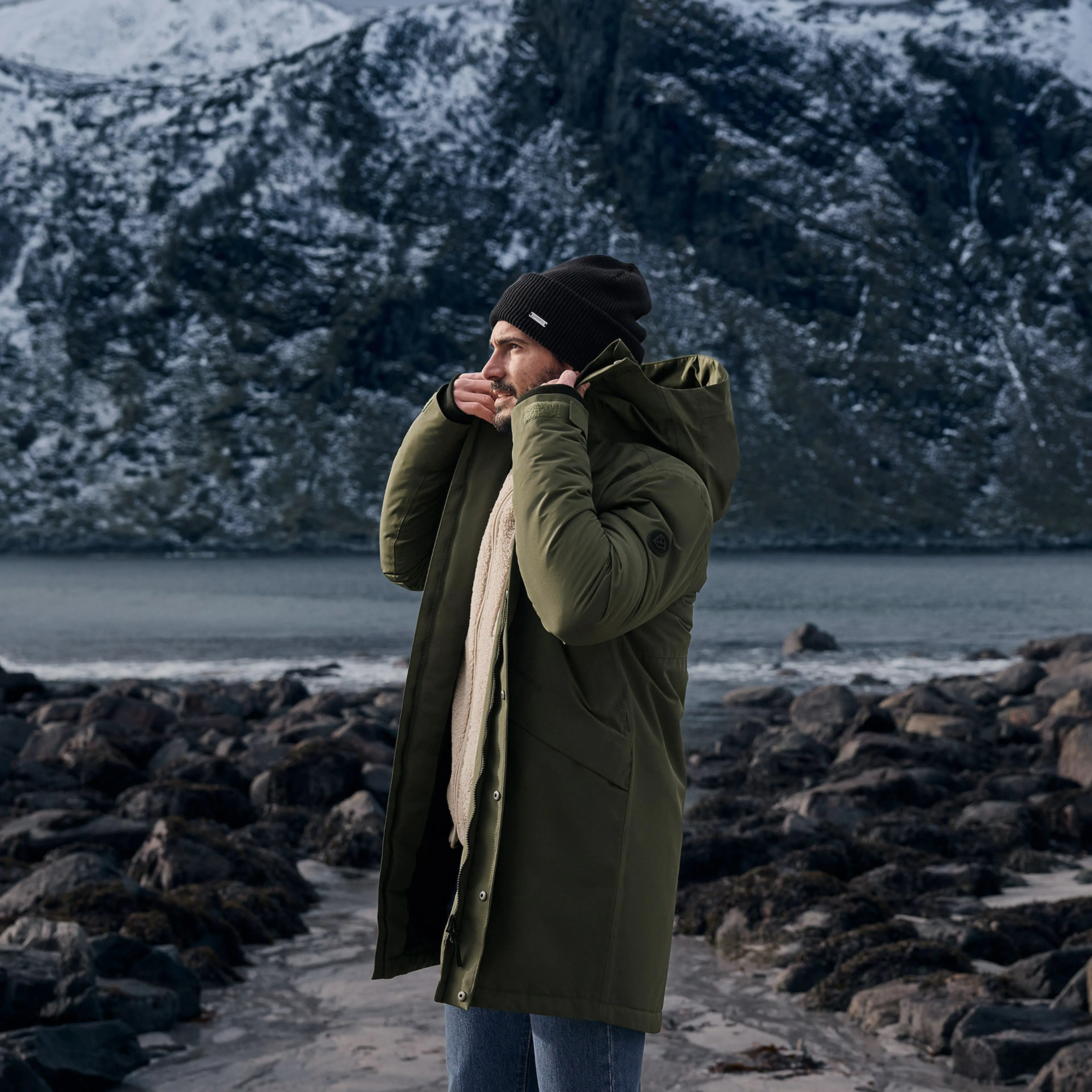 A person wearing a green coat and black beanie stands on a rocky shore with snow-capped mountains and a lake in the background.