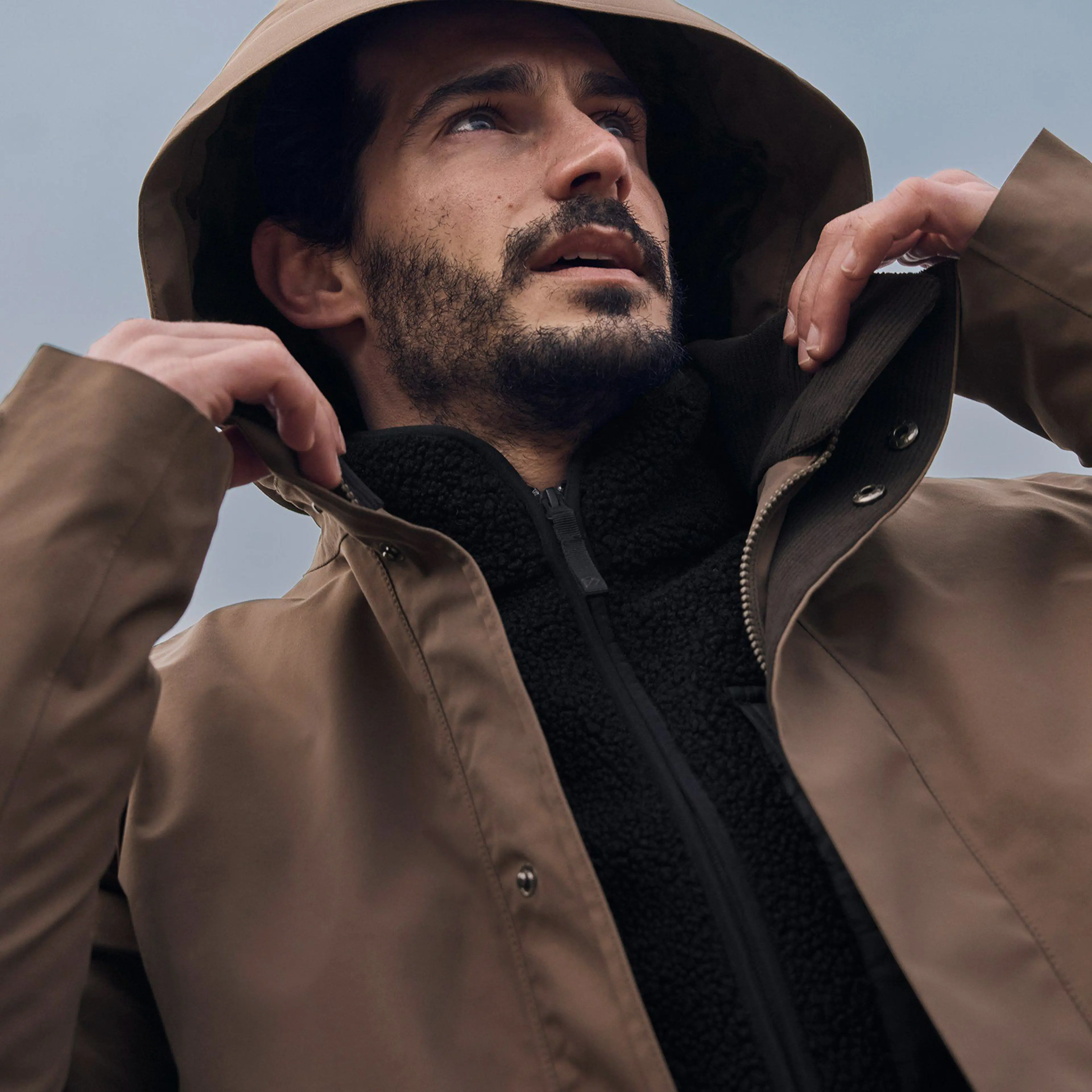 A man wearing a brown hooded jacket looks upward, adjusting his hood, against a cloudy sky background.