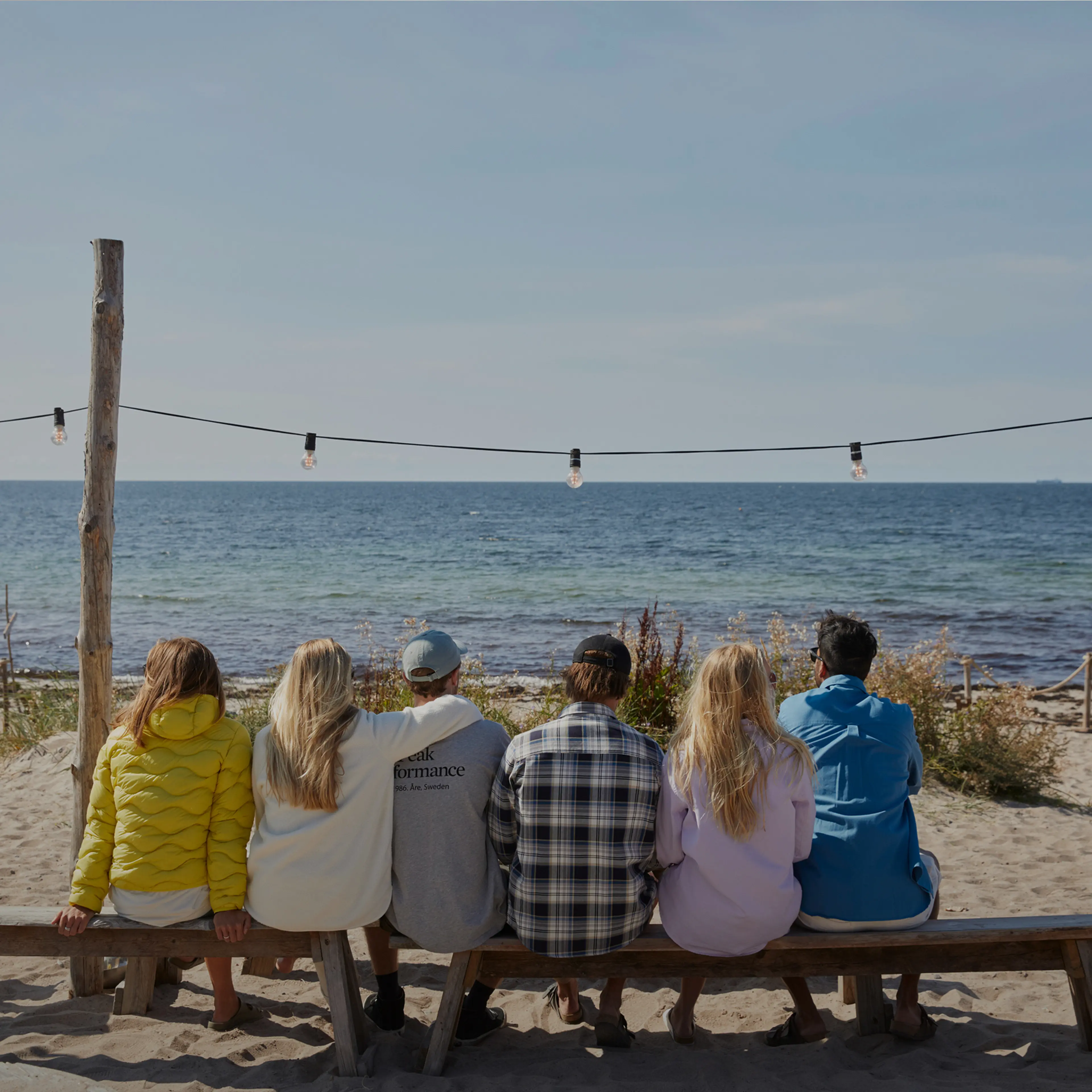 Five people sitting on a wooden bench at a sandy beach, looking at the ocean under a clear sky.