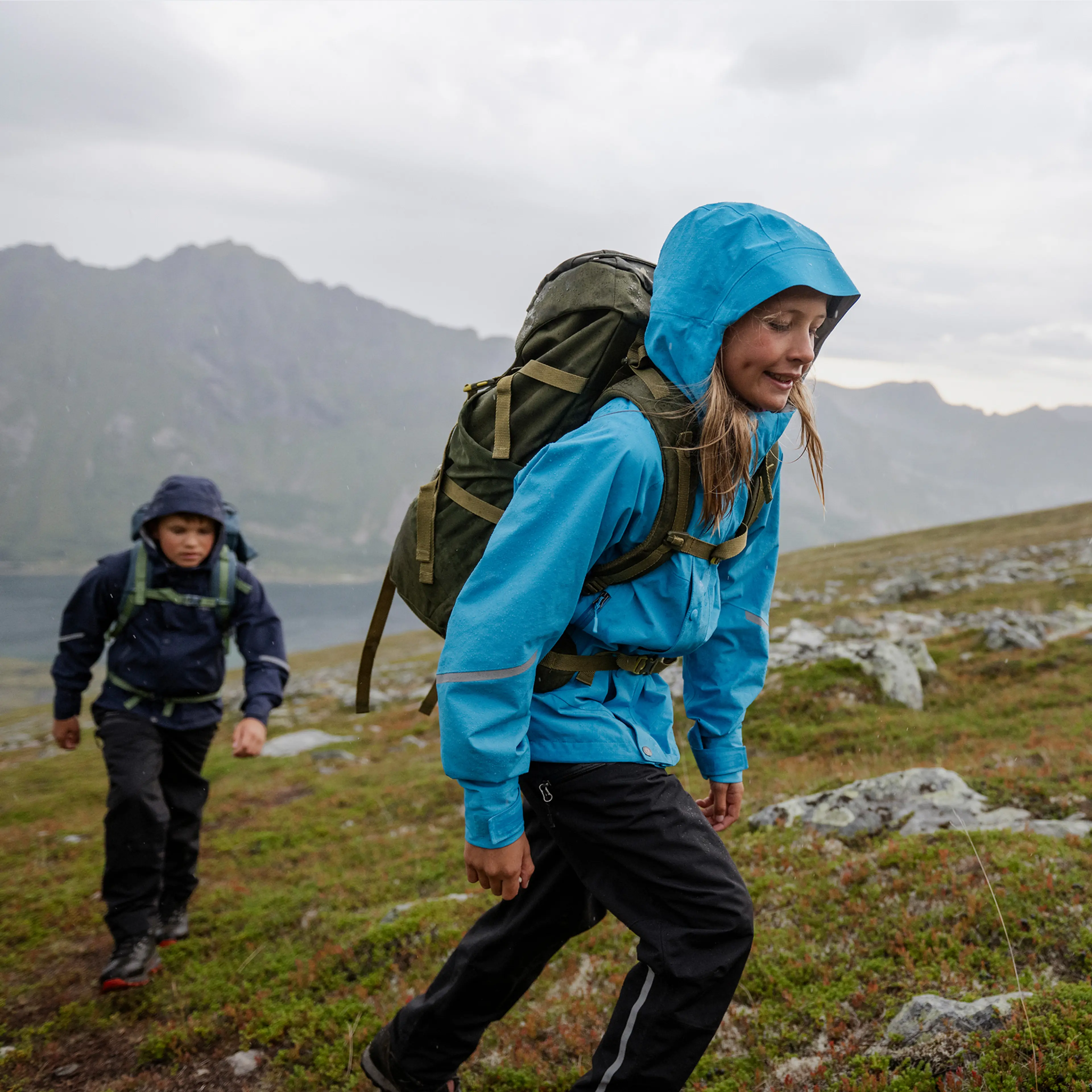 Two kids hiking uphill on a rocky path in rainy weather, wearing blue rain jackets with backpacks, against a backdrop of distant mountains.