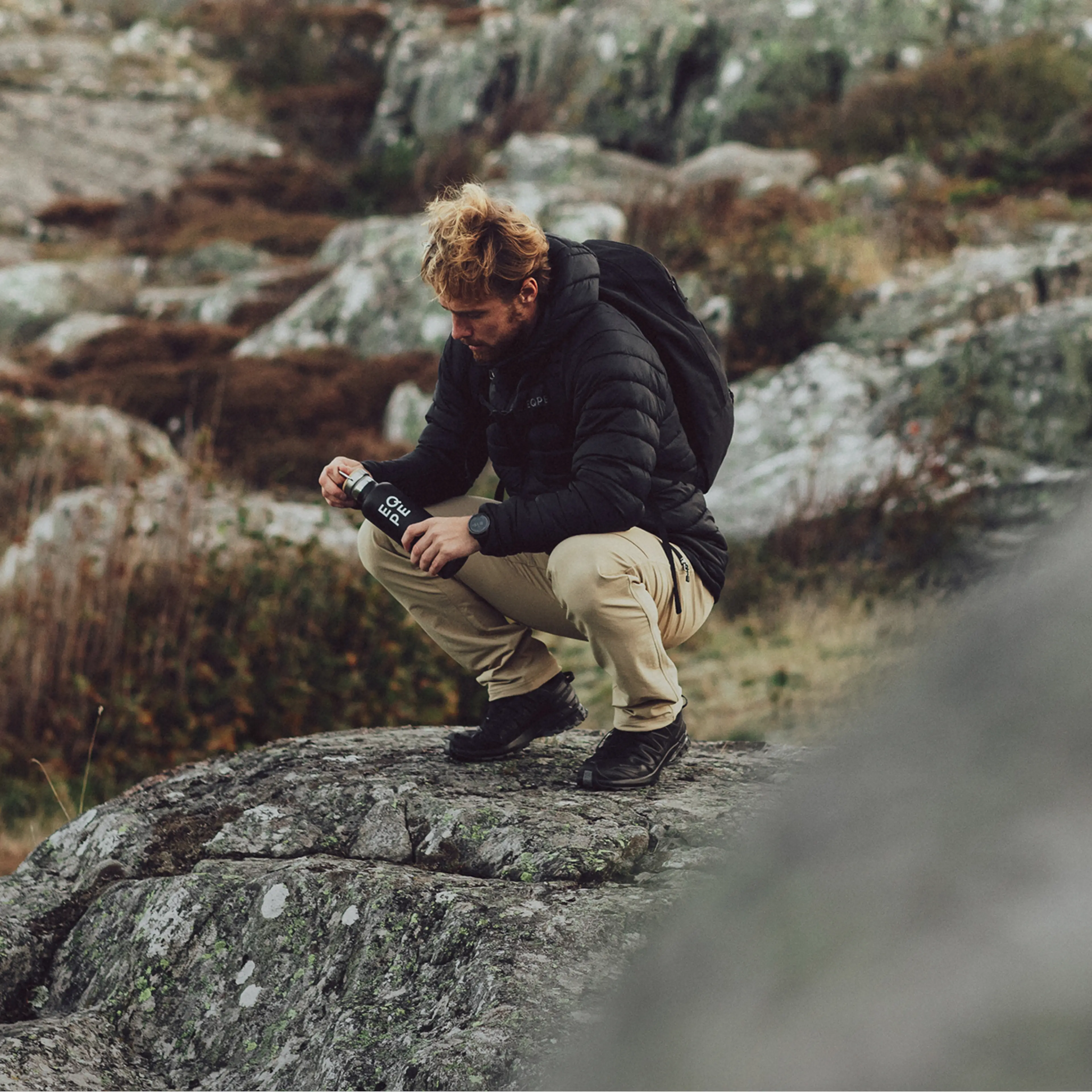 Man squatting on a rock, wearing a black jacket and beige pants, holding a can, with rocky terrain in the background.