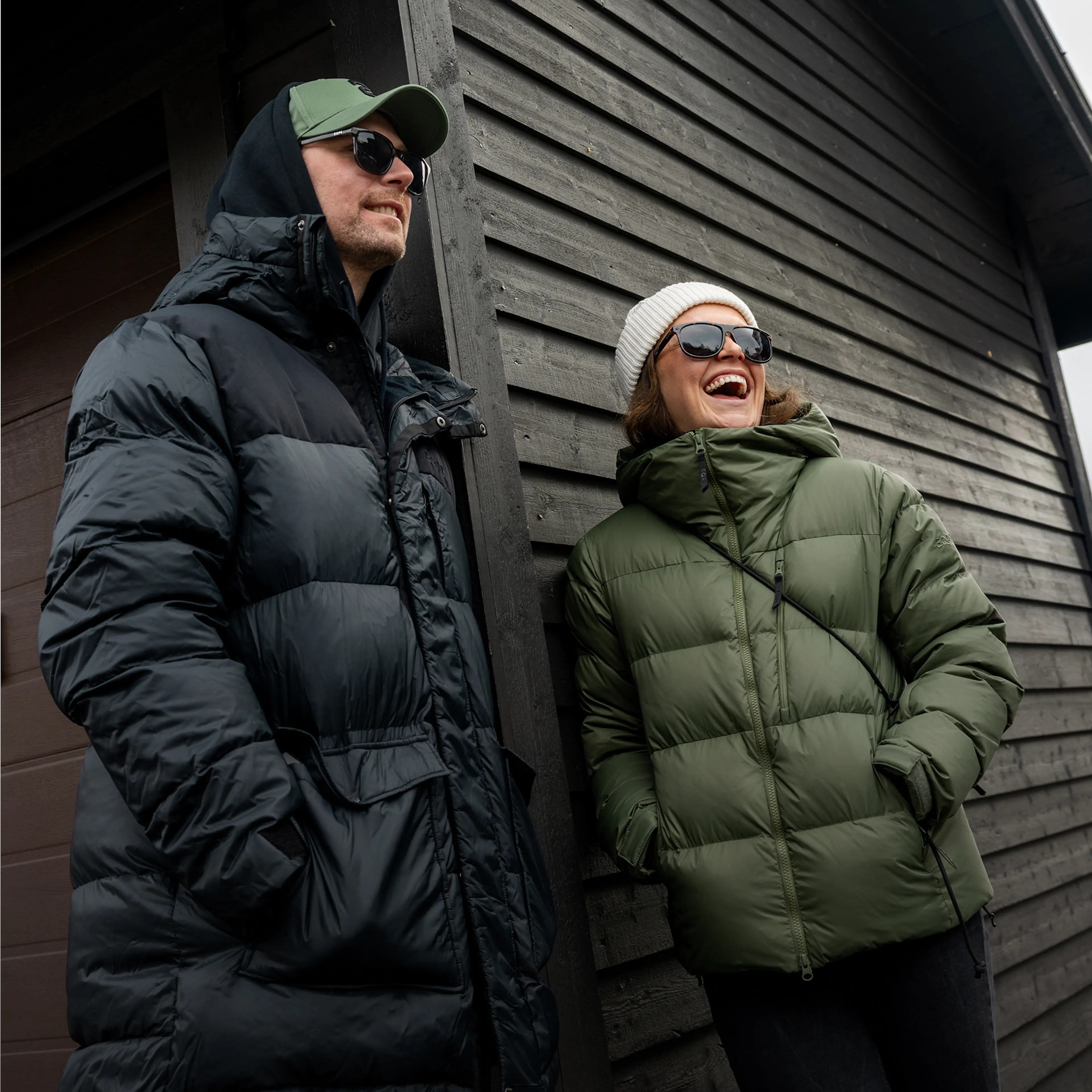 Two people in winter jackets and sunglasses smile and laugh as they stand against a dark wooden building.