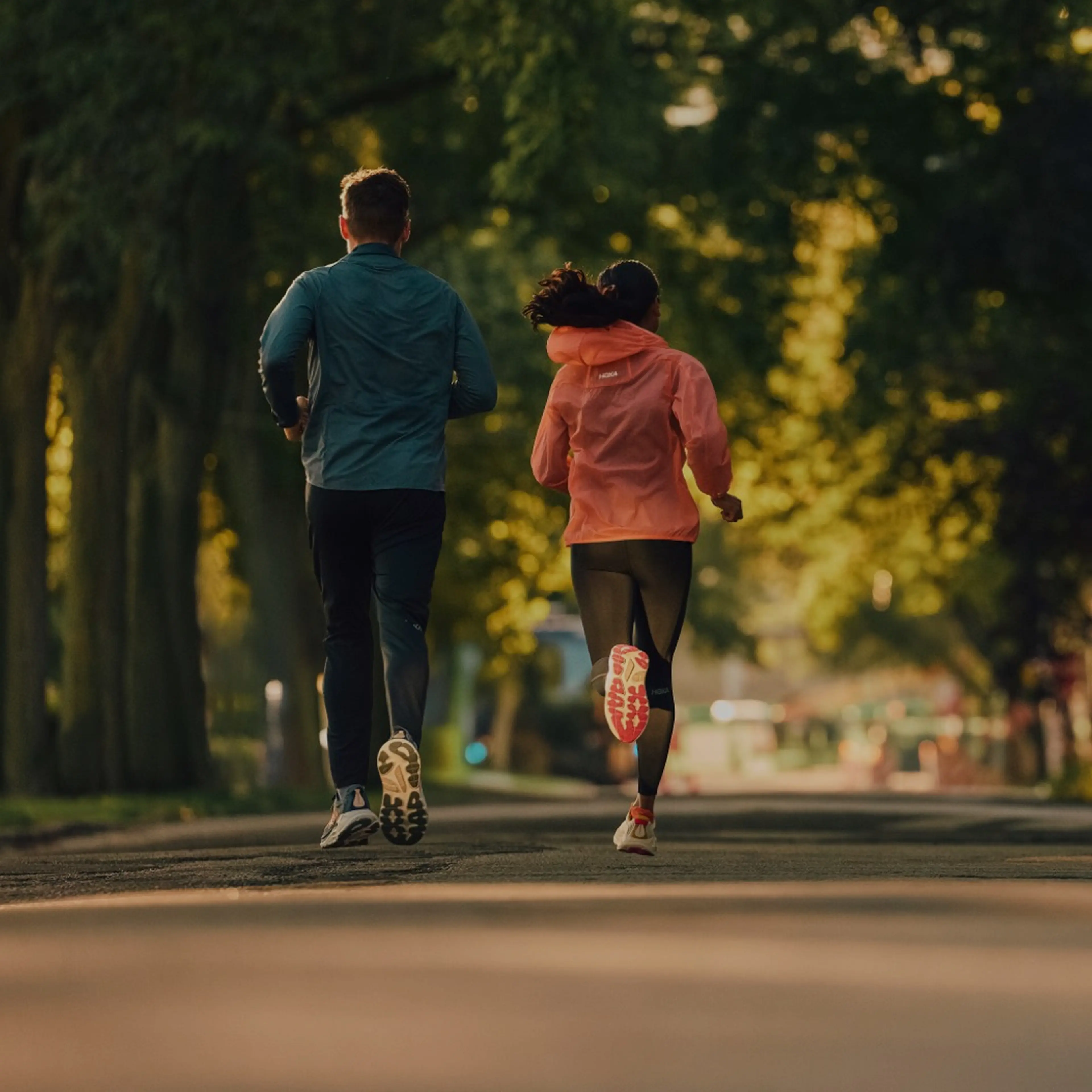 Two people jogging on a tree-lined path in the park, wearing athletic outfits. The sun filters through the leaves creating a serene ambiance.