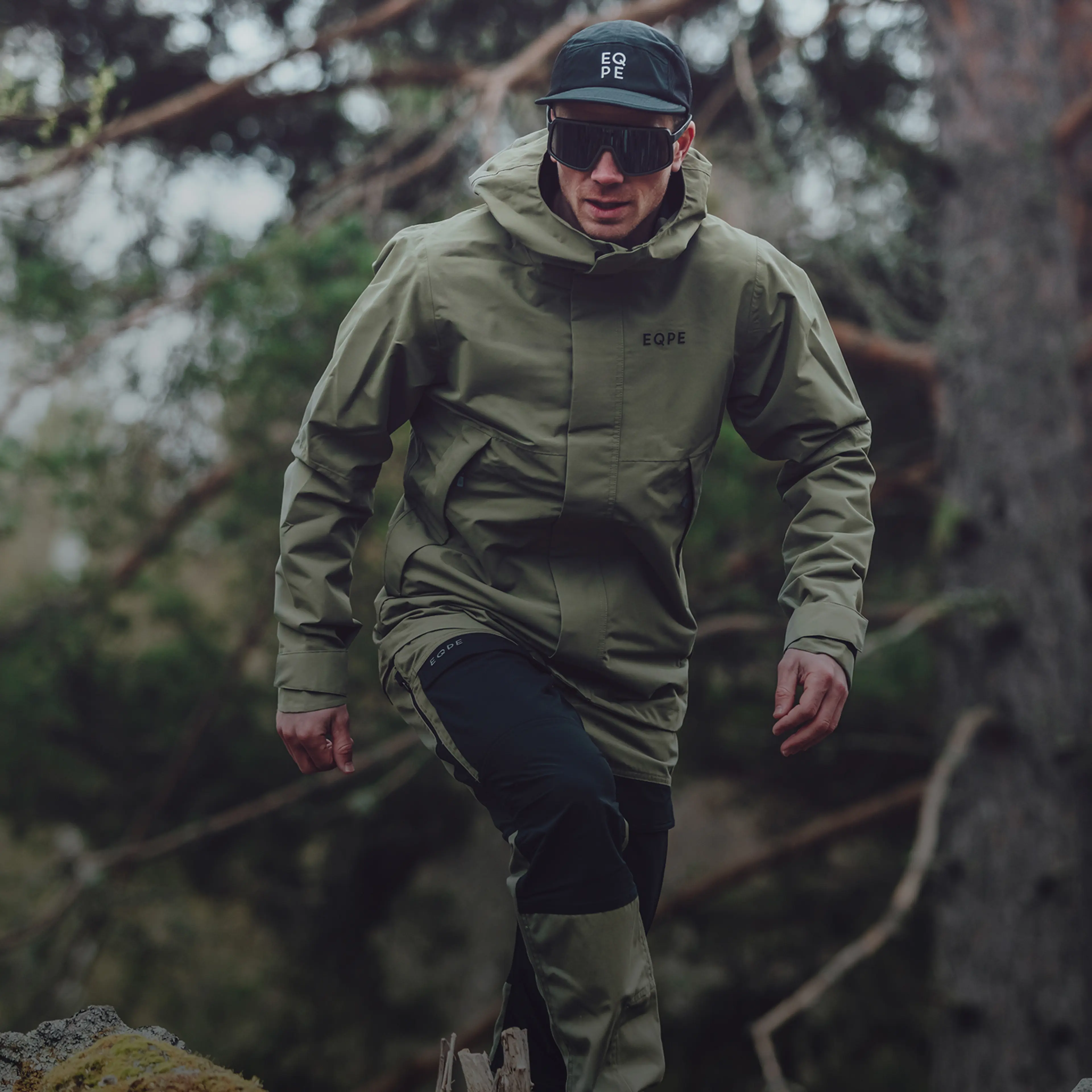 A man in outdoor gear, including a cap and sunglasses, hikes through a forested area, showcasing a rugged and dynamic pose.