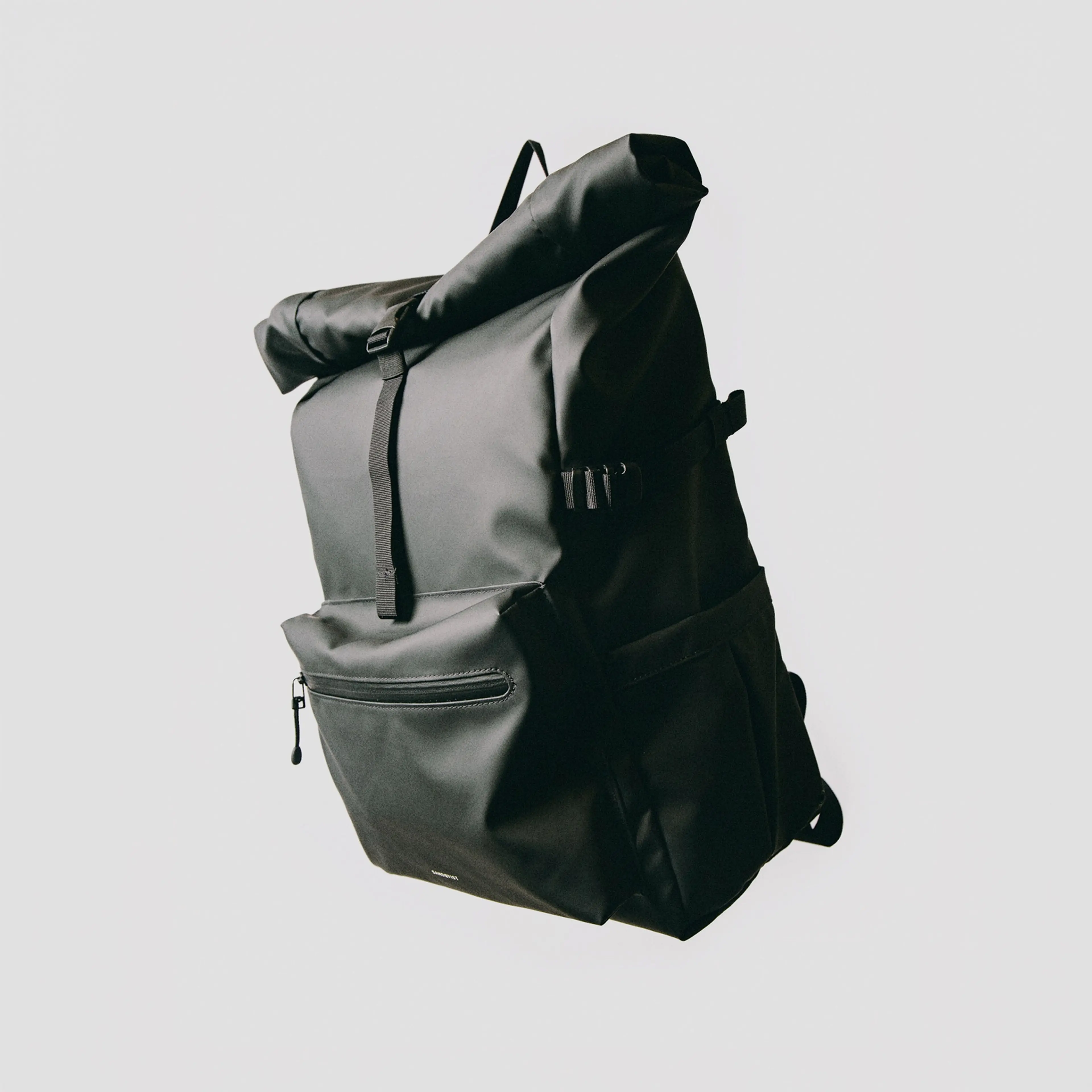 Black roll-top backpack with a front zipper pocket and side buckles, set against a plain white background.