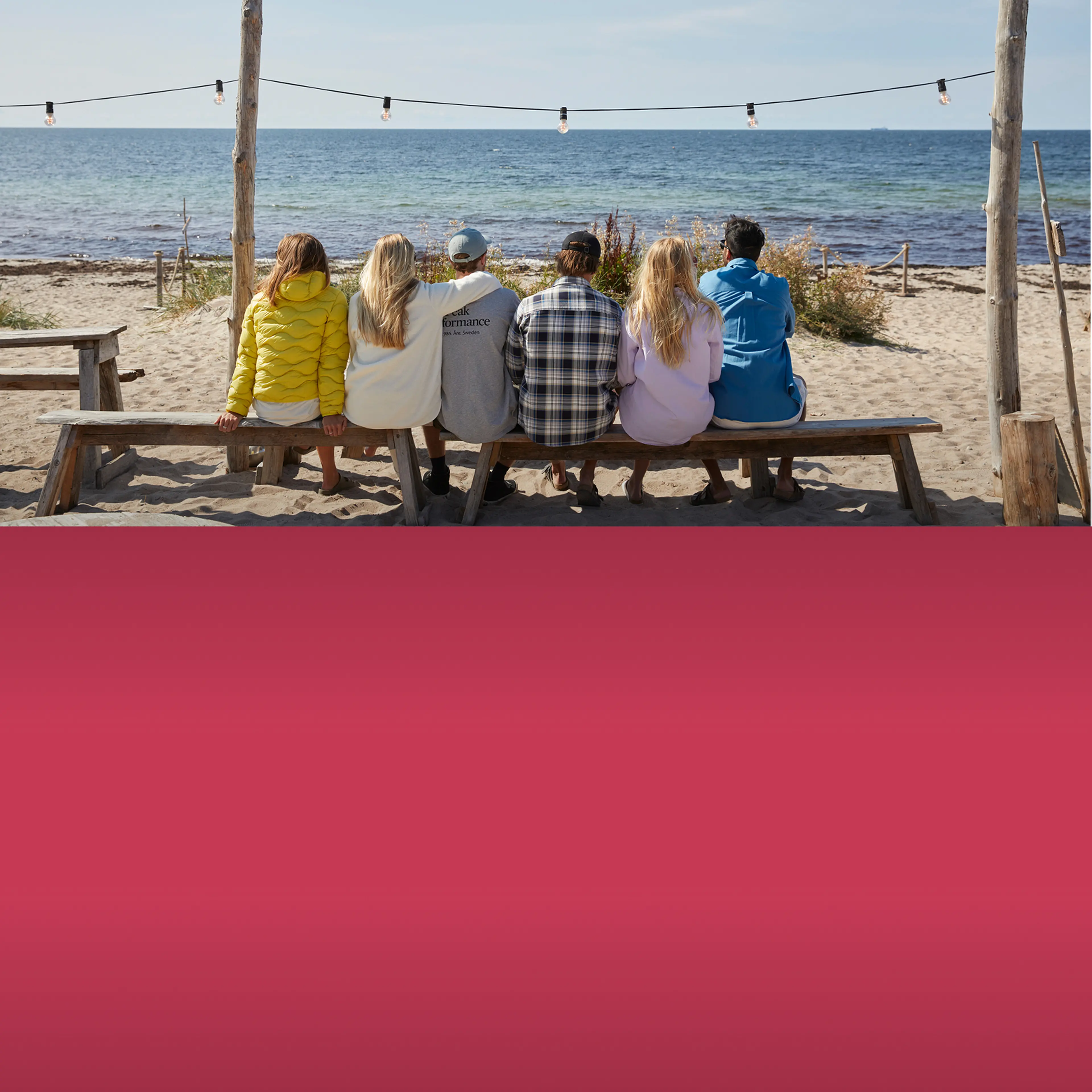 Six people sit on a beach bench facing the sea, under hanging string lights, with a sandy shore and clear sky in the background.