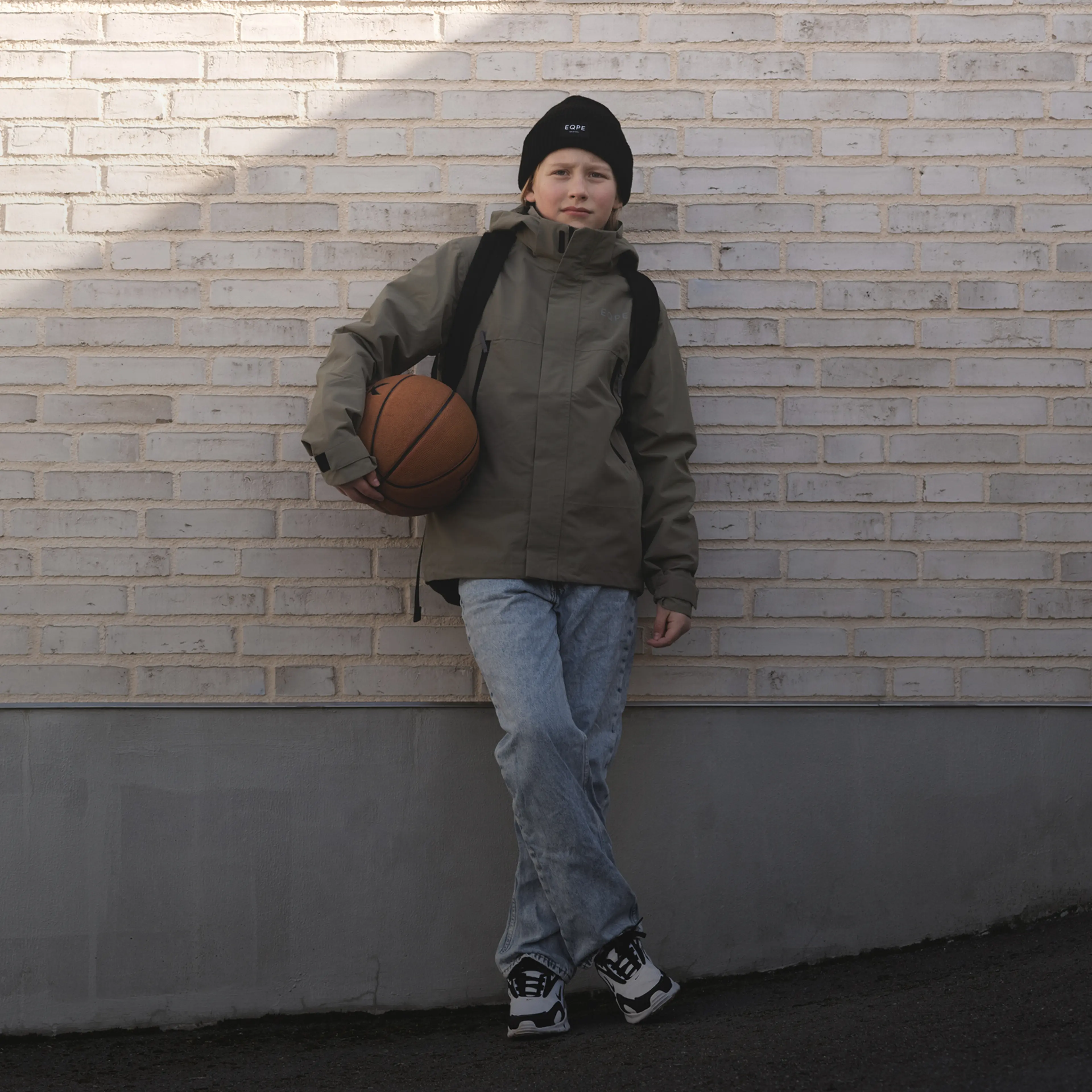 A person wearing a green jacket, jeans, and a black beanie leans against a brick wall, holding a basketball under their arm.