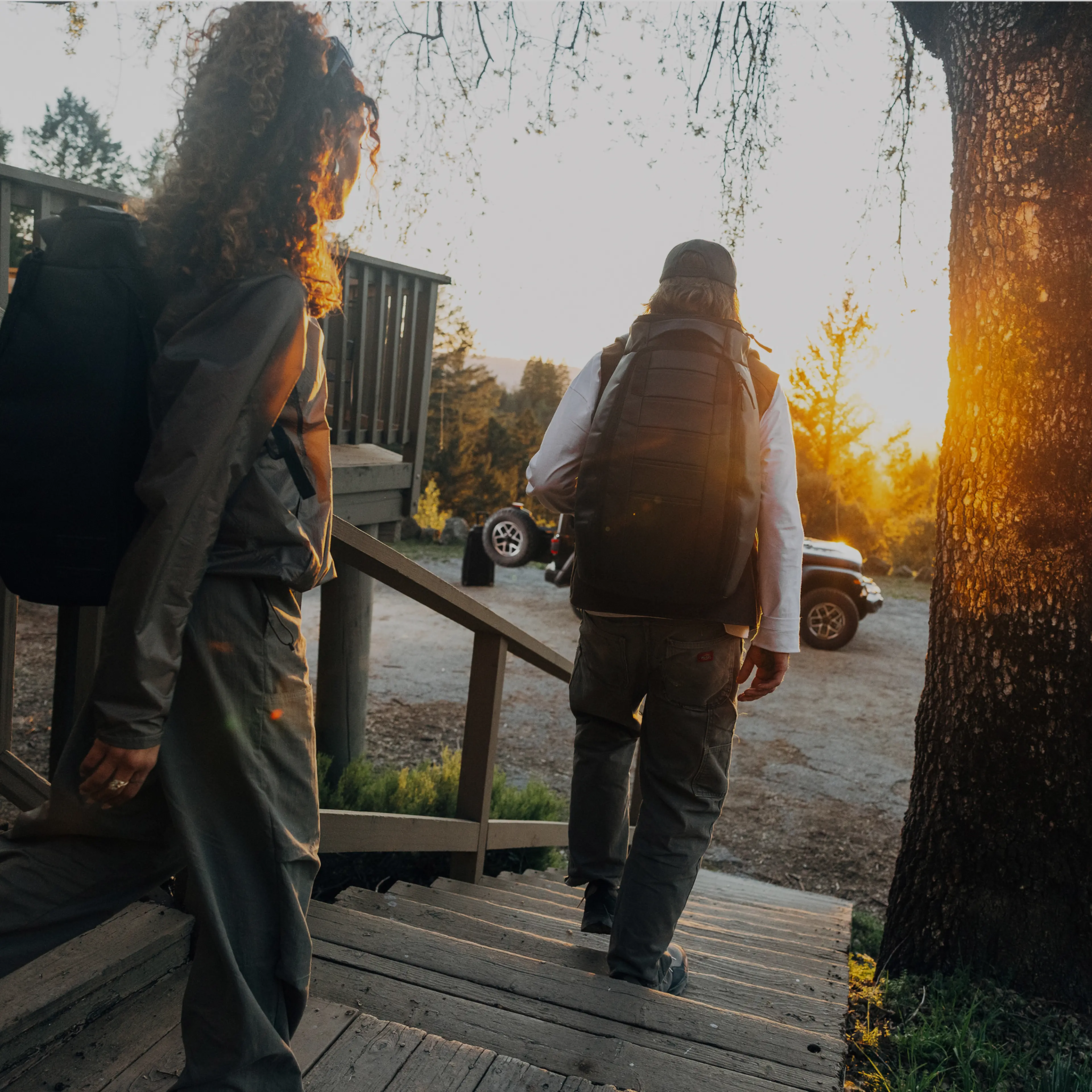 Two people with backpacks walk down wooden steps, approaching a car at sunset, surrounded by trees and a forested landscape.