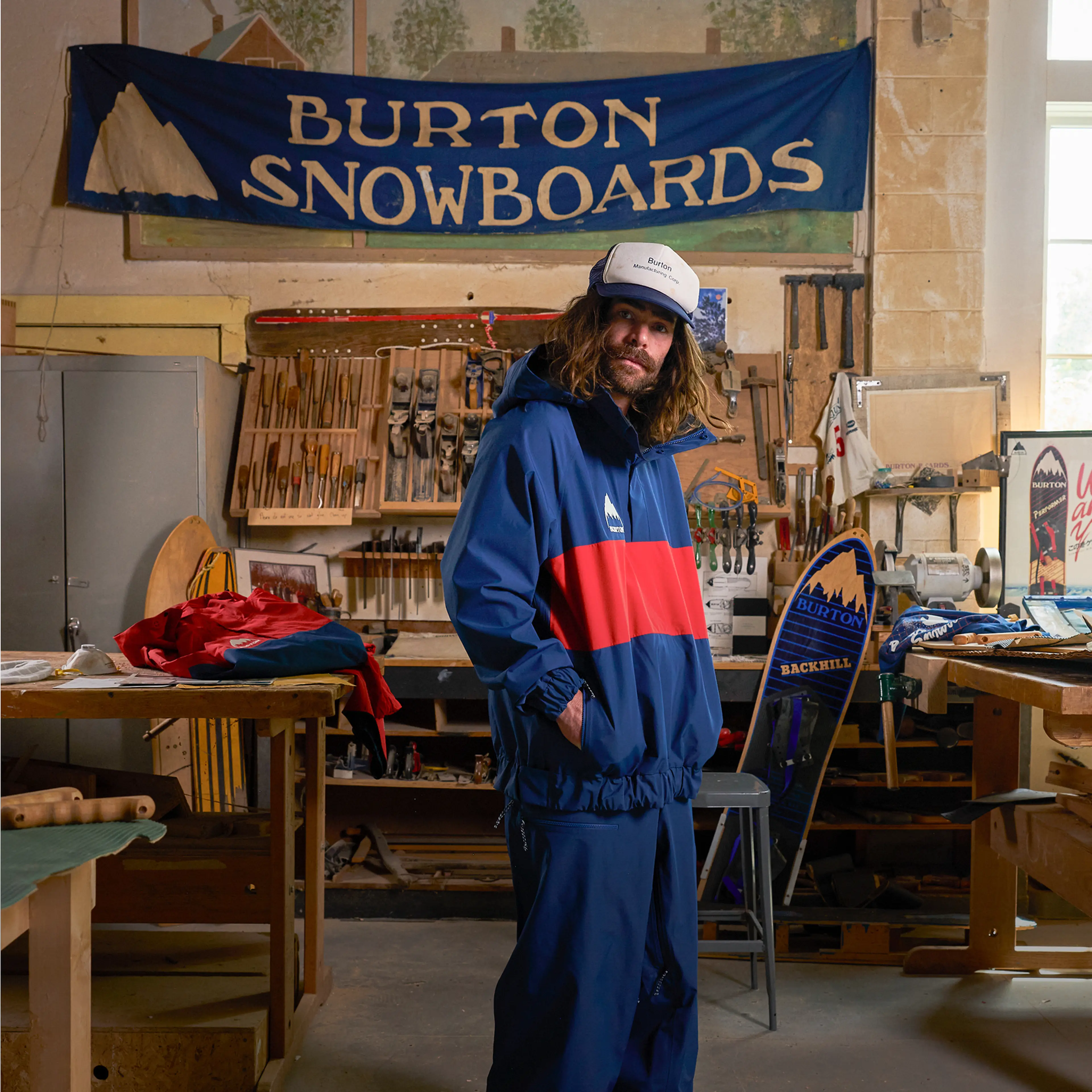 A person in a blue and red jacket stands in a workshop, surrounded by tools and snowboards, under a "Burton Snowboards" banner.