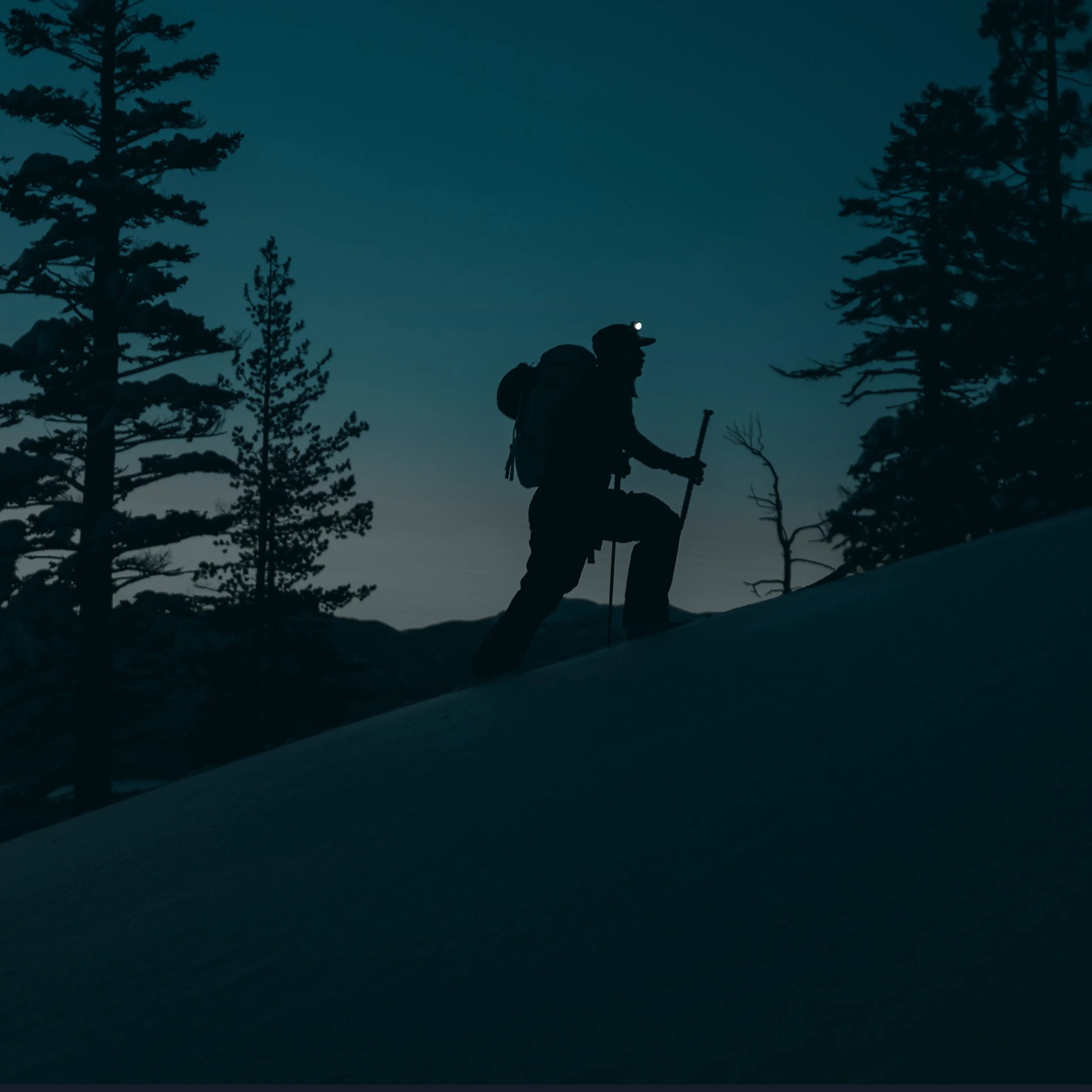 Silhouette of a hiker with a headlamp and backpack, climbing a snowy hill at dusk, surrounded by trees.