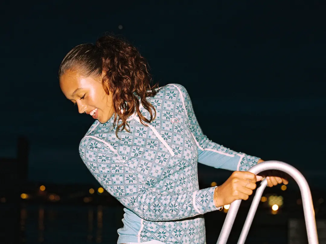 Woman in a patterned sweater climbing a ladder at night, smiling with city lights glowing in the background.