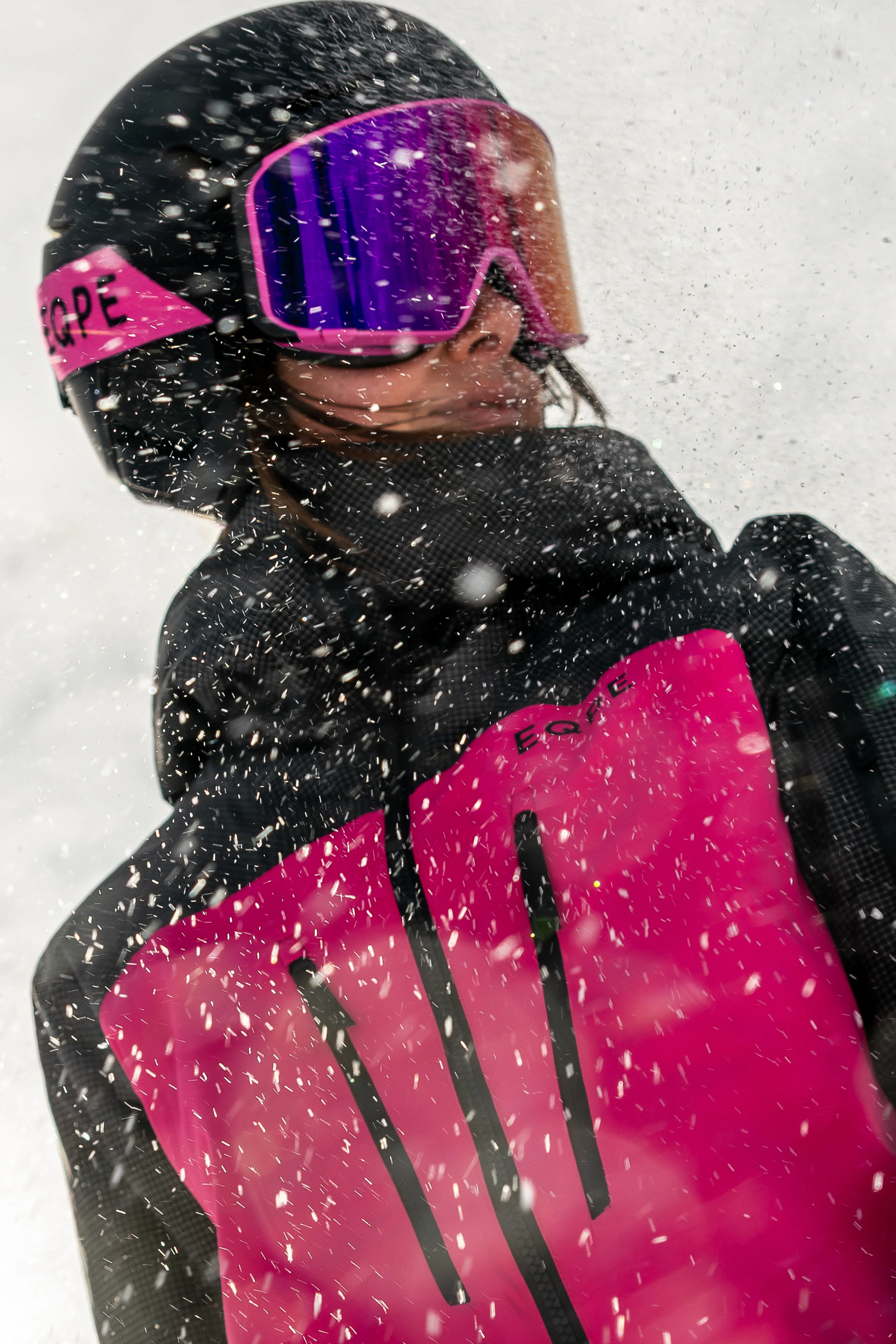 A person wearing a pink and black snowboard jacket, helmet, and reflective goggles, surrounded by falling snow.