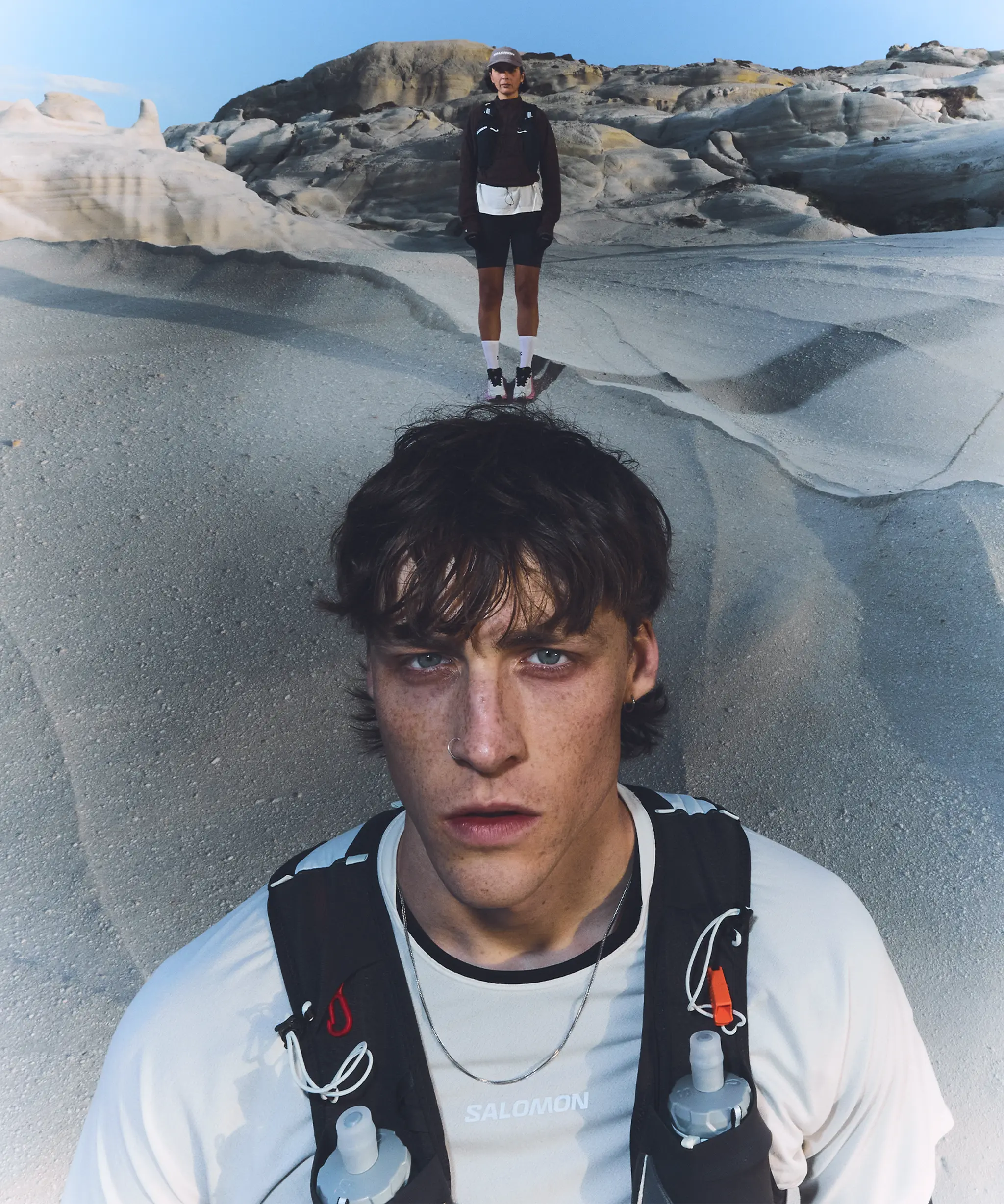 Two people wearing athletic gear stand on a rocky, sandy terrain under a clear blue sky. The focus is on the person in the foreground.