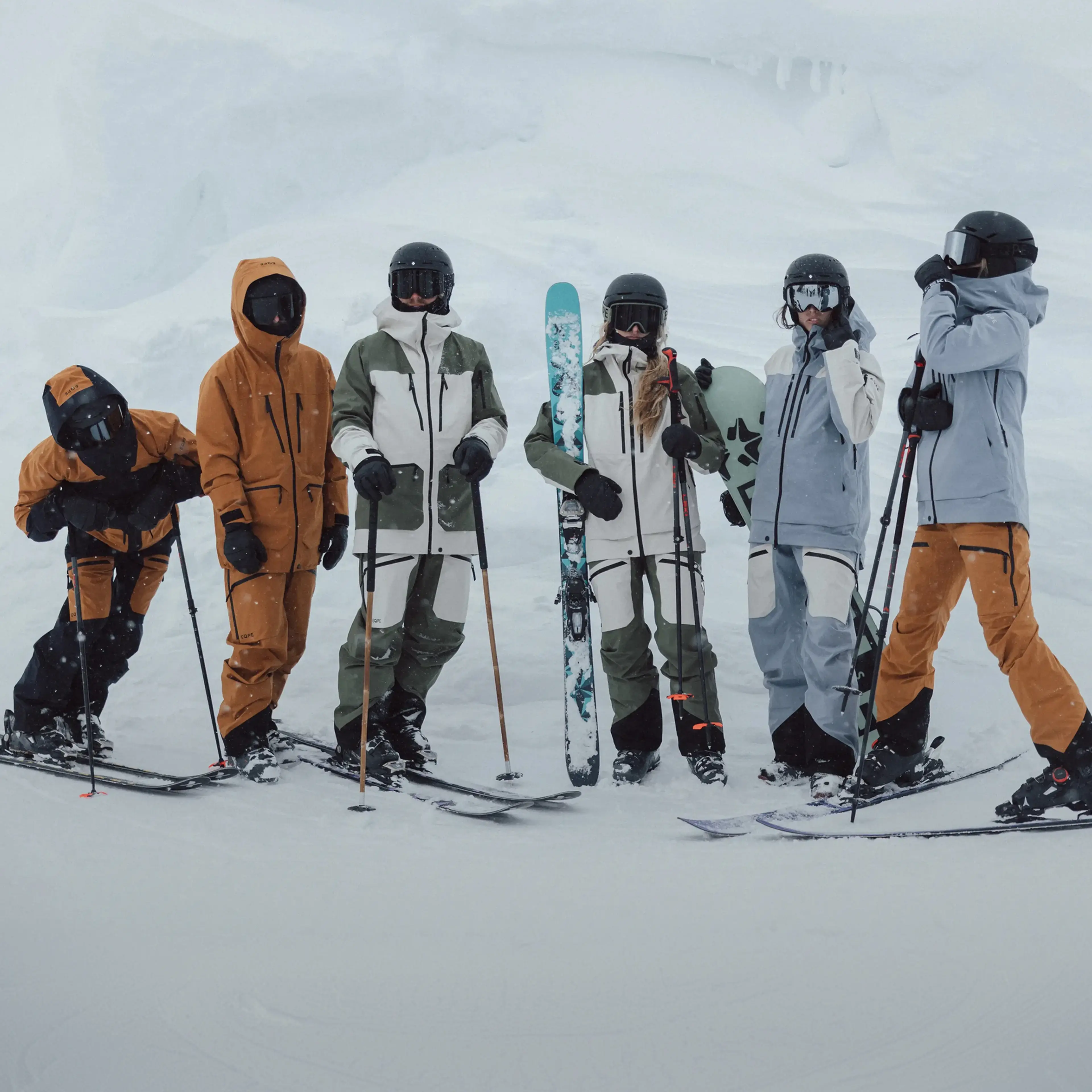 A group of six skiers in winter gear standing together on a snowy slope, holding skis and poles, with snowboards visible.