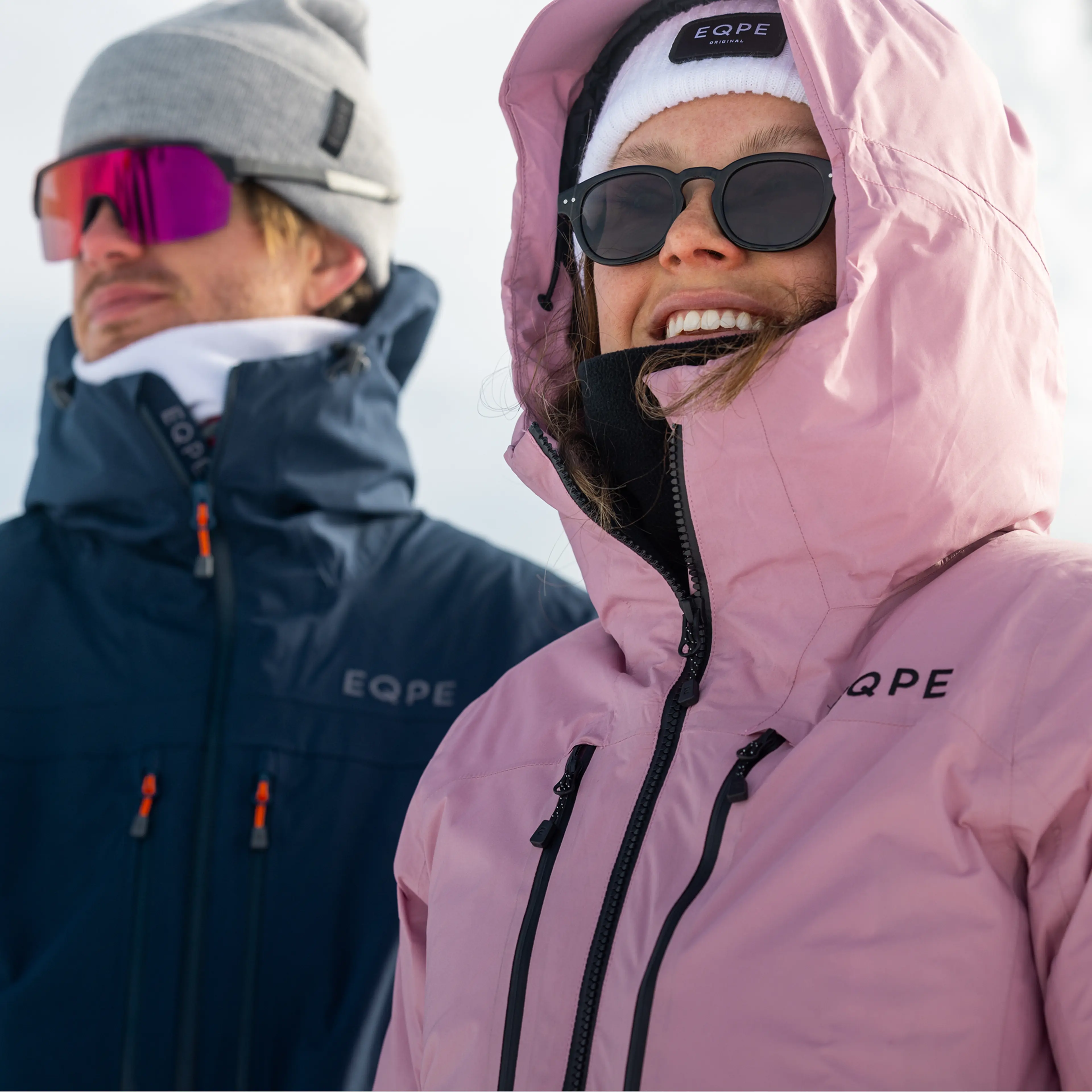 Two people in winter jackets and sunglasses, smiling on a snowy day. One wears a pink coat, the other a blue one with a gray beanie.