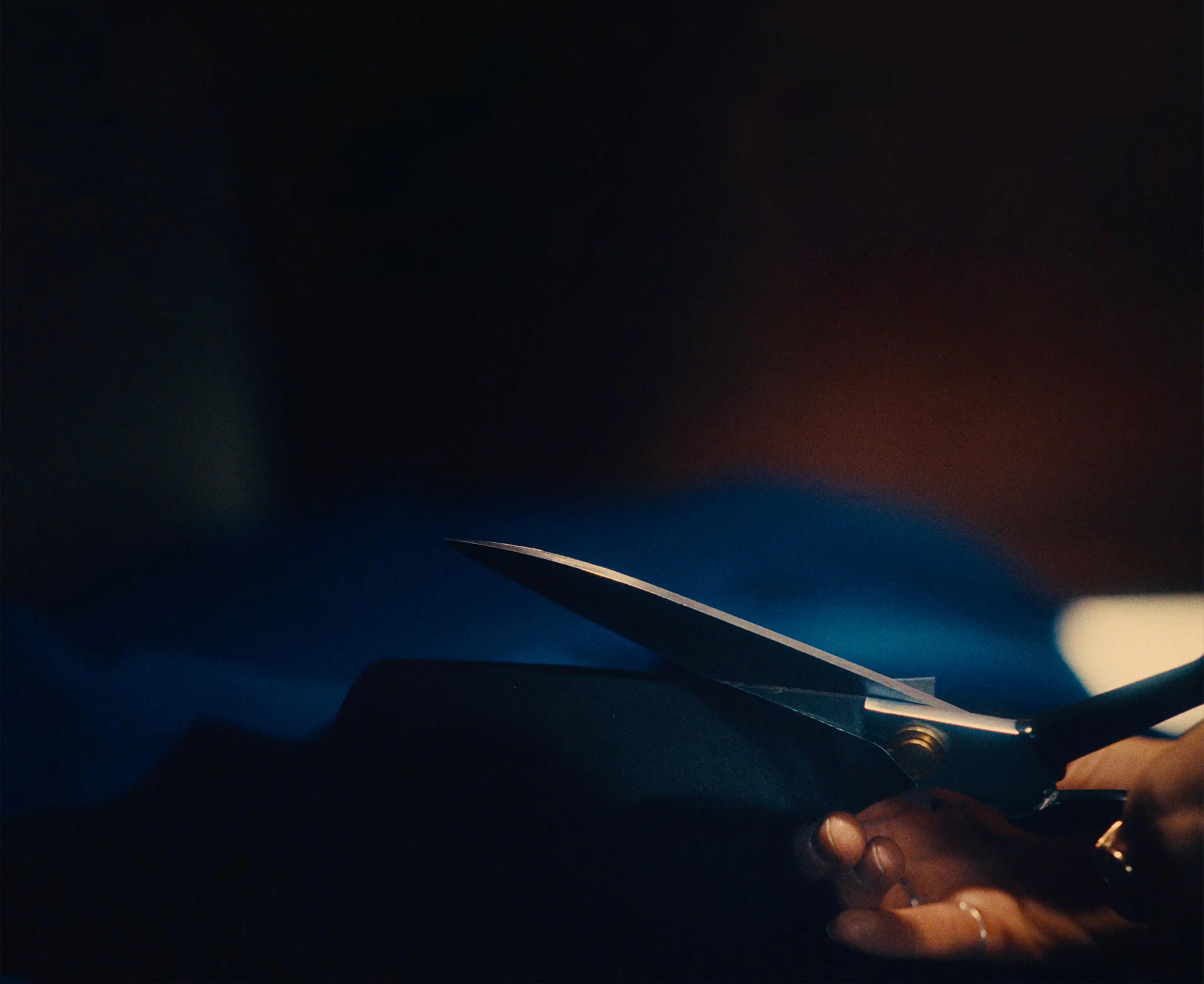 Sara's hand holding black scissors, cutting through dark fabric in dim lighting