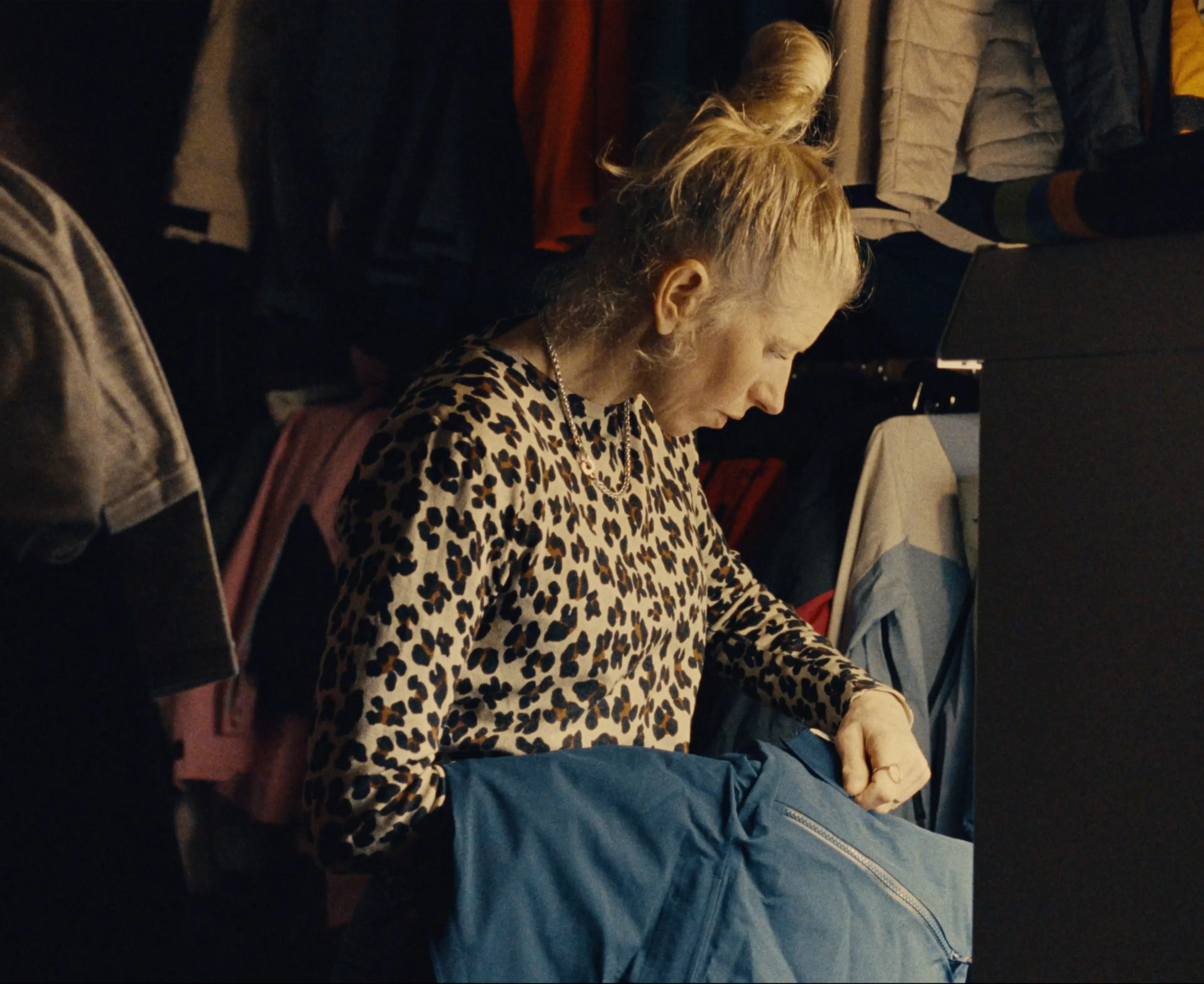 Sara examining a blue garment in a dimly lit room filled with clothes