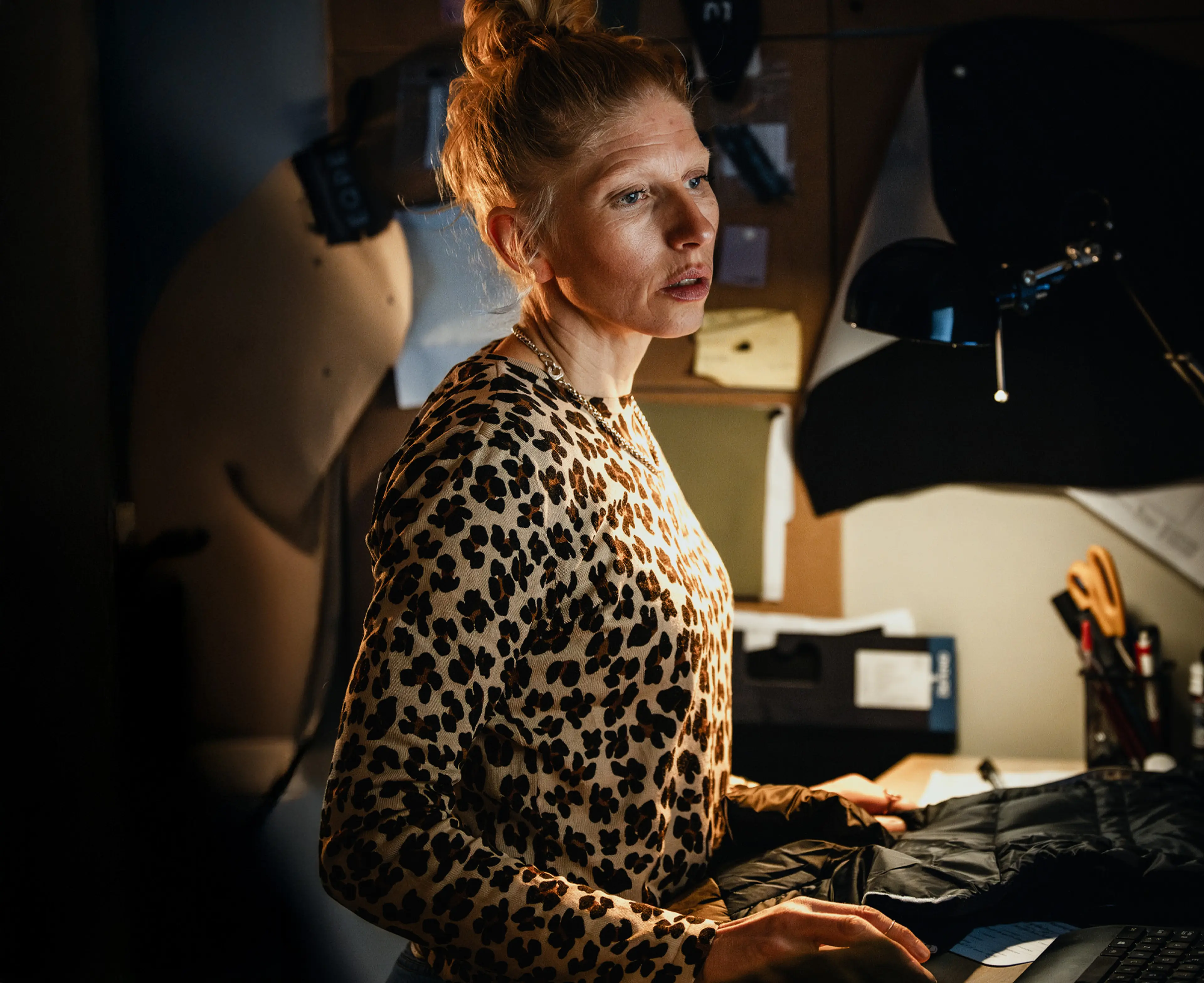 Sara stands focused in a dimly lit workspace, surrounded by sewing materials and patterns on the wall.