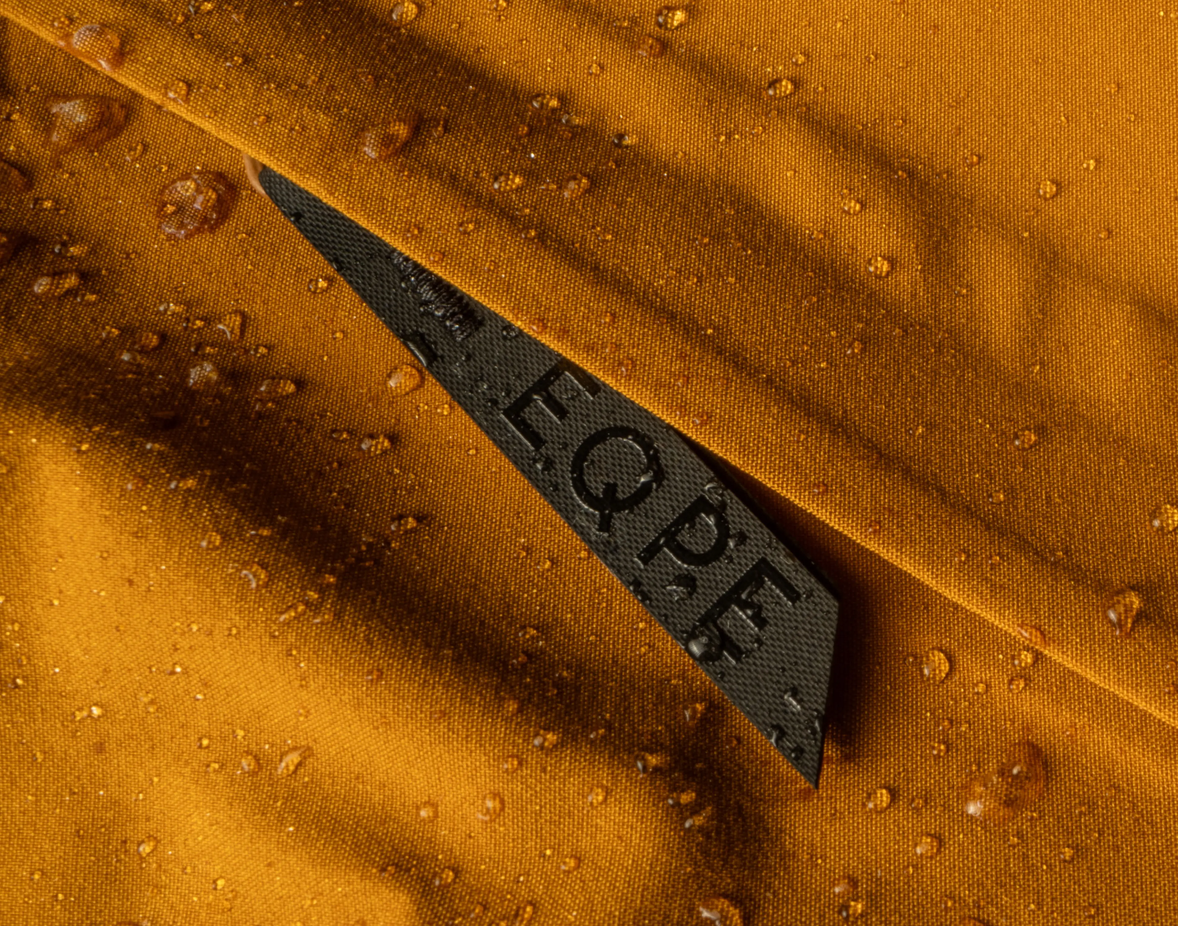 Close-up of water droplets on orange waterproof fabric with a black label partially visible, featuring the letters "EQPE."