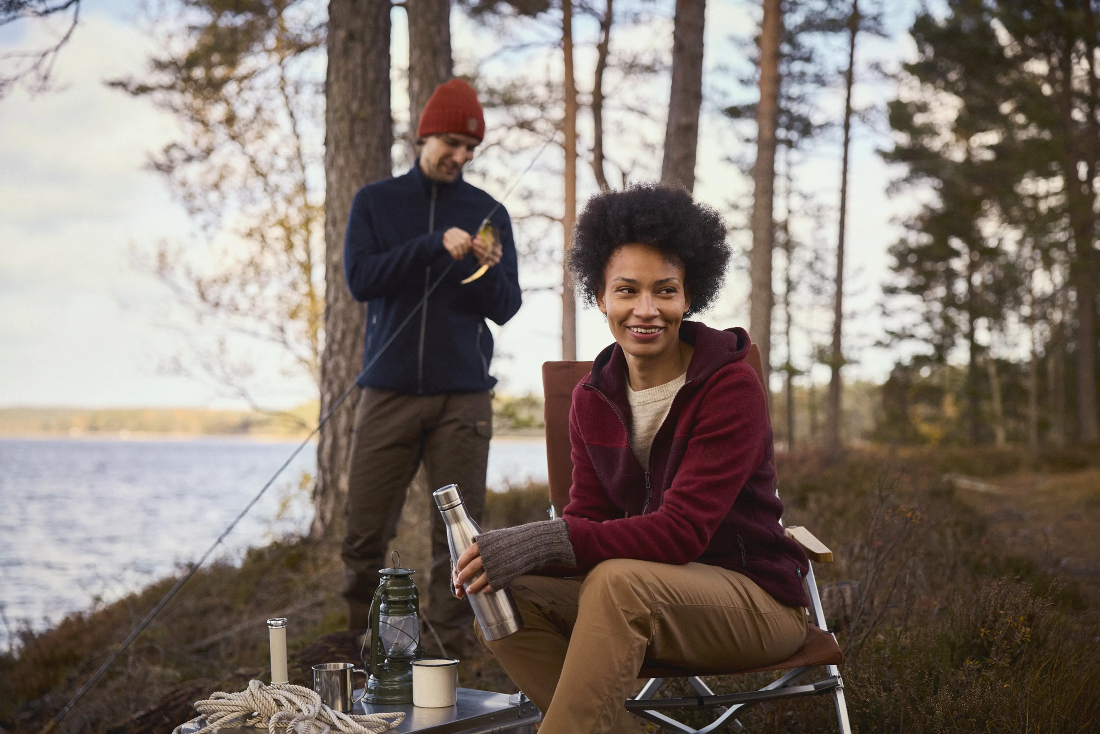 A woman sits smiling with a thermos near a lake, while a man, wearing a red beanie, prepares a fishing rod in a wooded area.