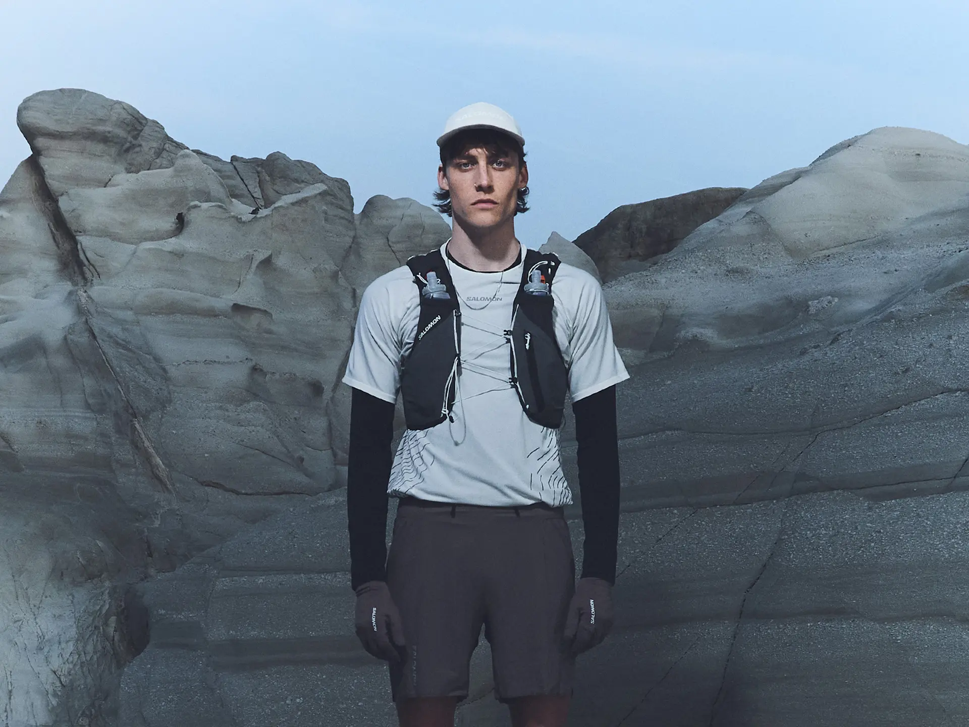 A person wearing outdoor sports gear and a hydration vest stands in front of rugged rock formations under a clear sky.