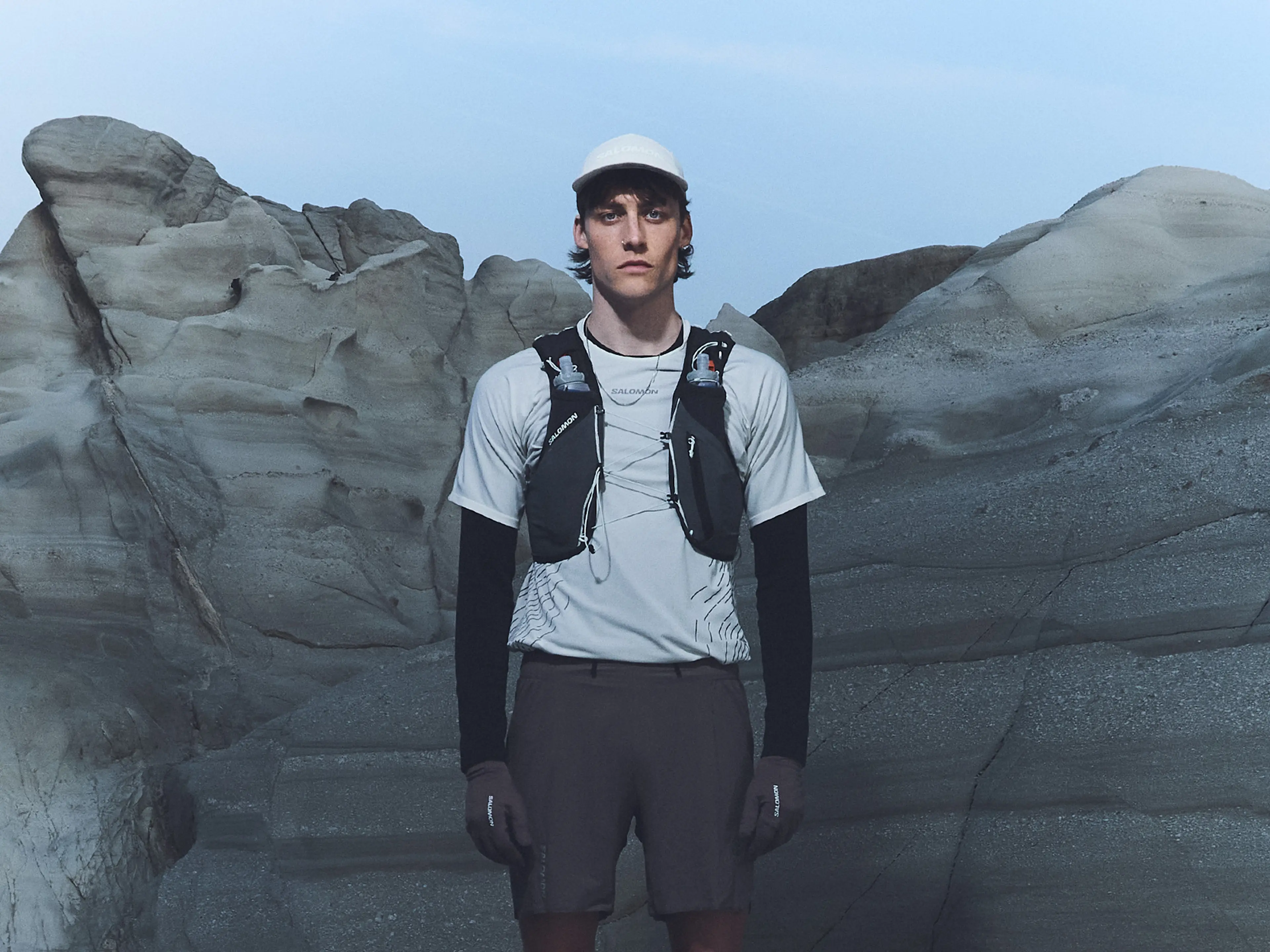 A person wearing outdoor sports gear and a hydration vest stands in front of rugged rock formations under a clear sky.