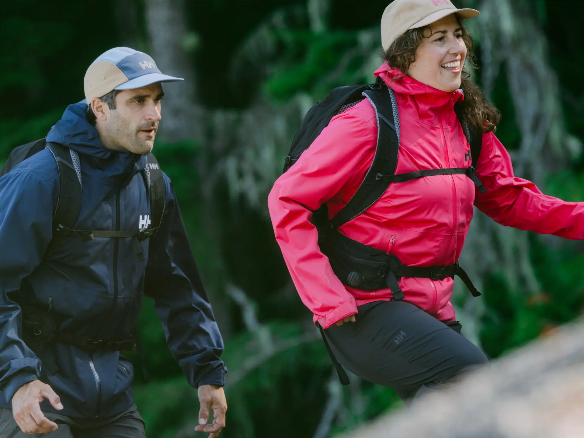 Two hikers in colorful jackets and caps are trekking through a forest, smiling and enjoying the outdoor adventure.