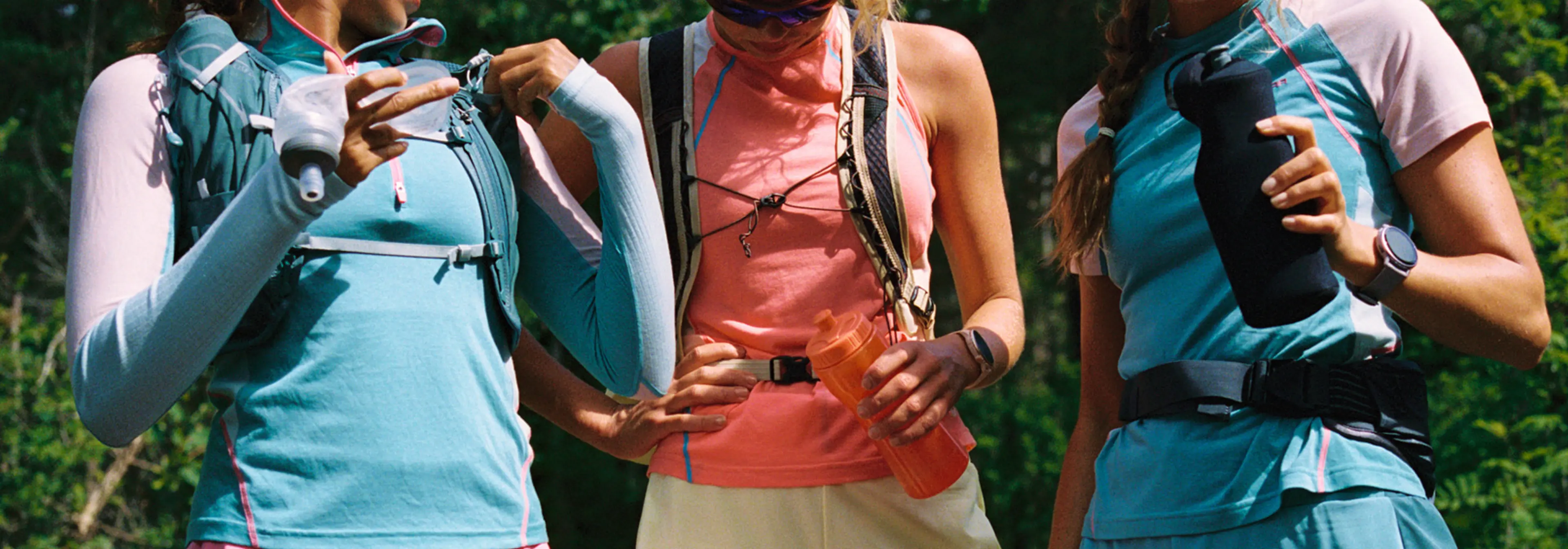 Three women in athletic gear and hydration packs prepare for a run, holding water bottles amid a lush outdoor setting.