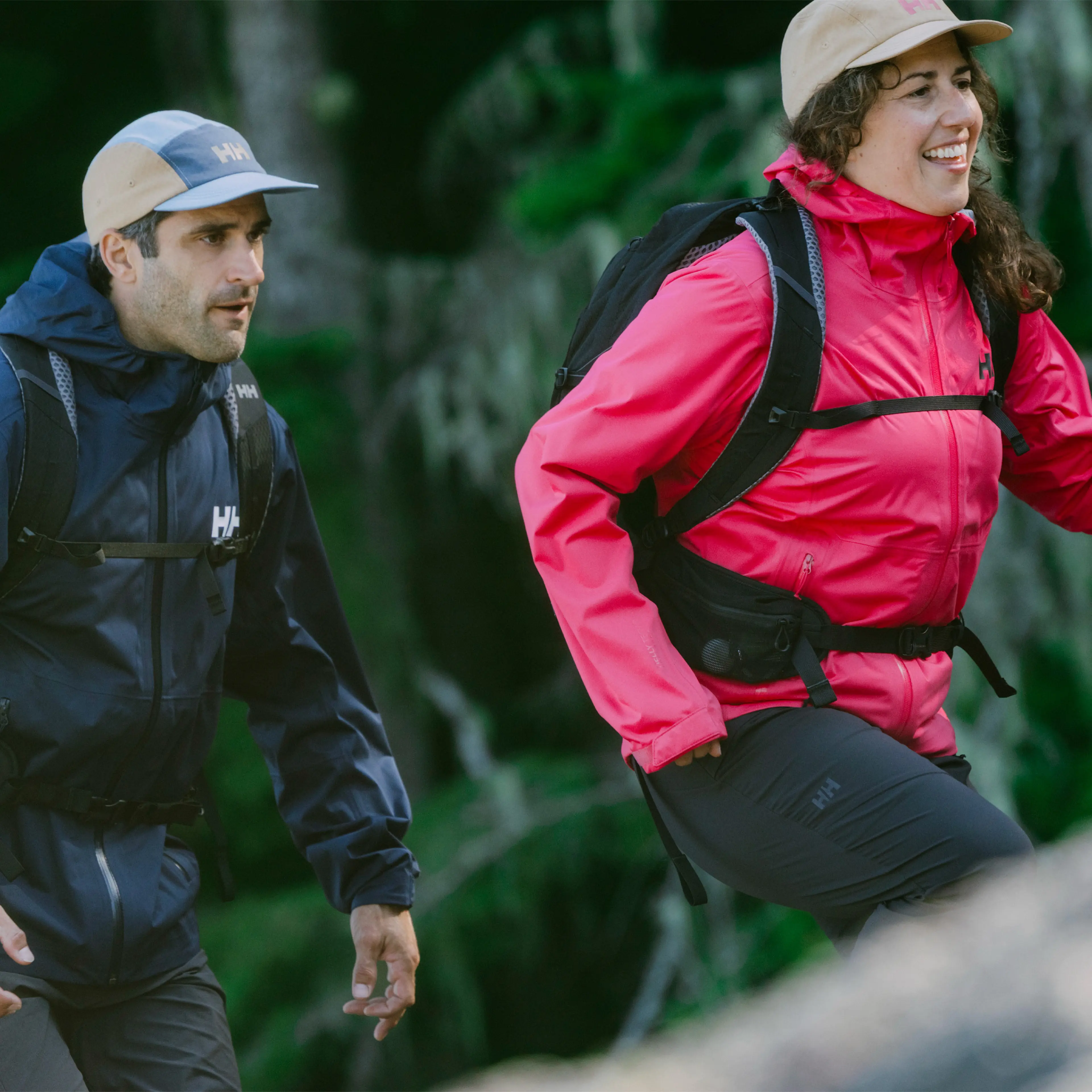 Two people hiking in the forest, wearing caps, jackets, and backpacks; one in pink and the other in navy blue.