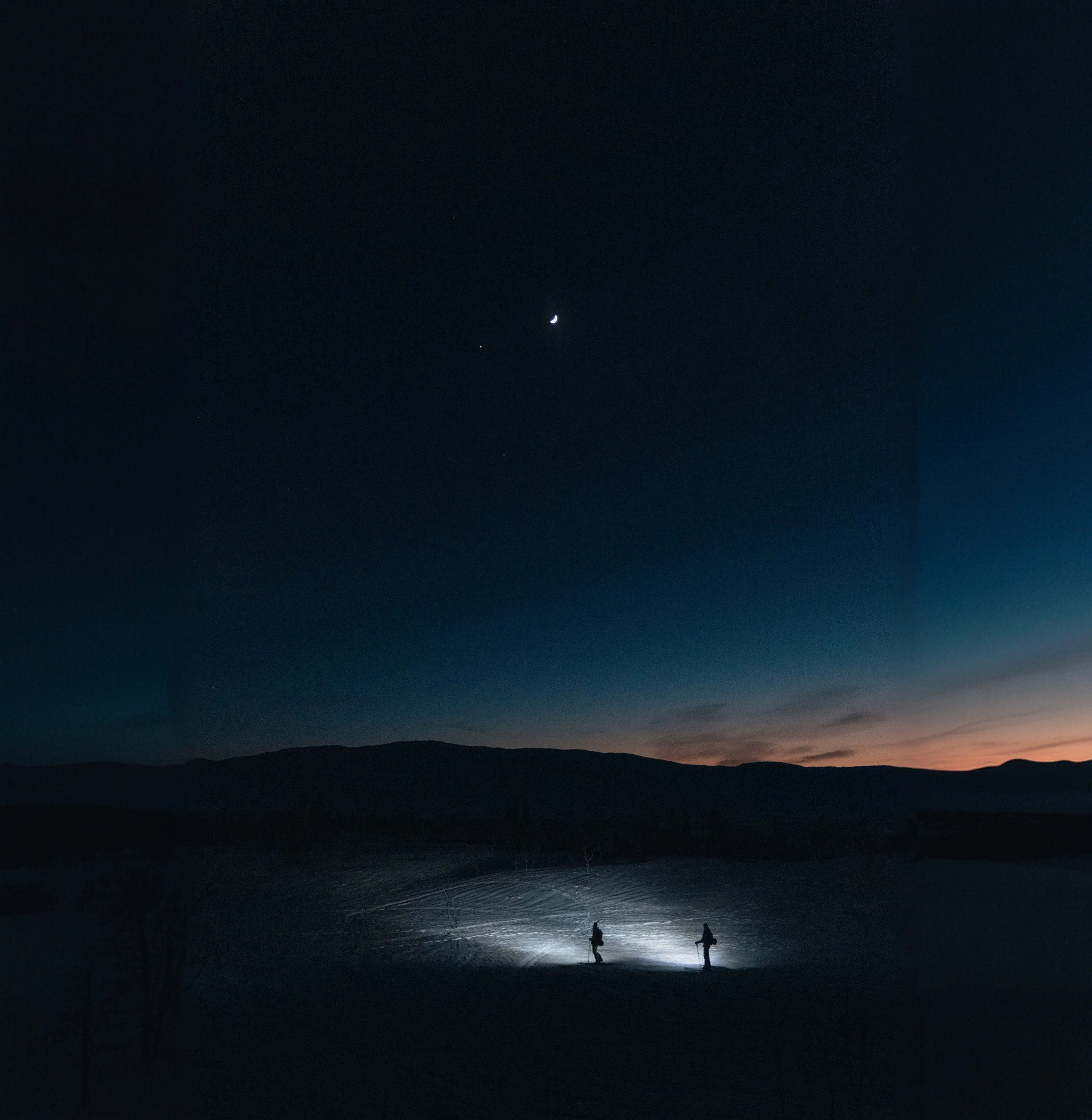 Two people with flashlights stand on snow under a crescent moon and stars against a dark mountain silhouette and a fading sunset.