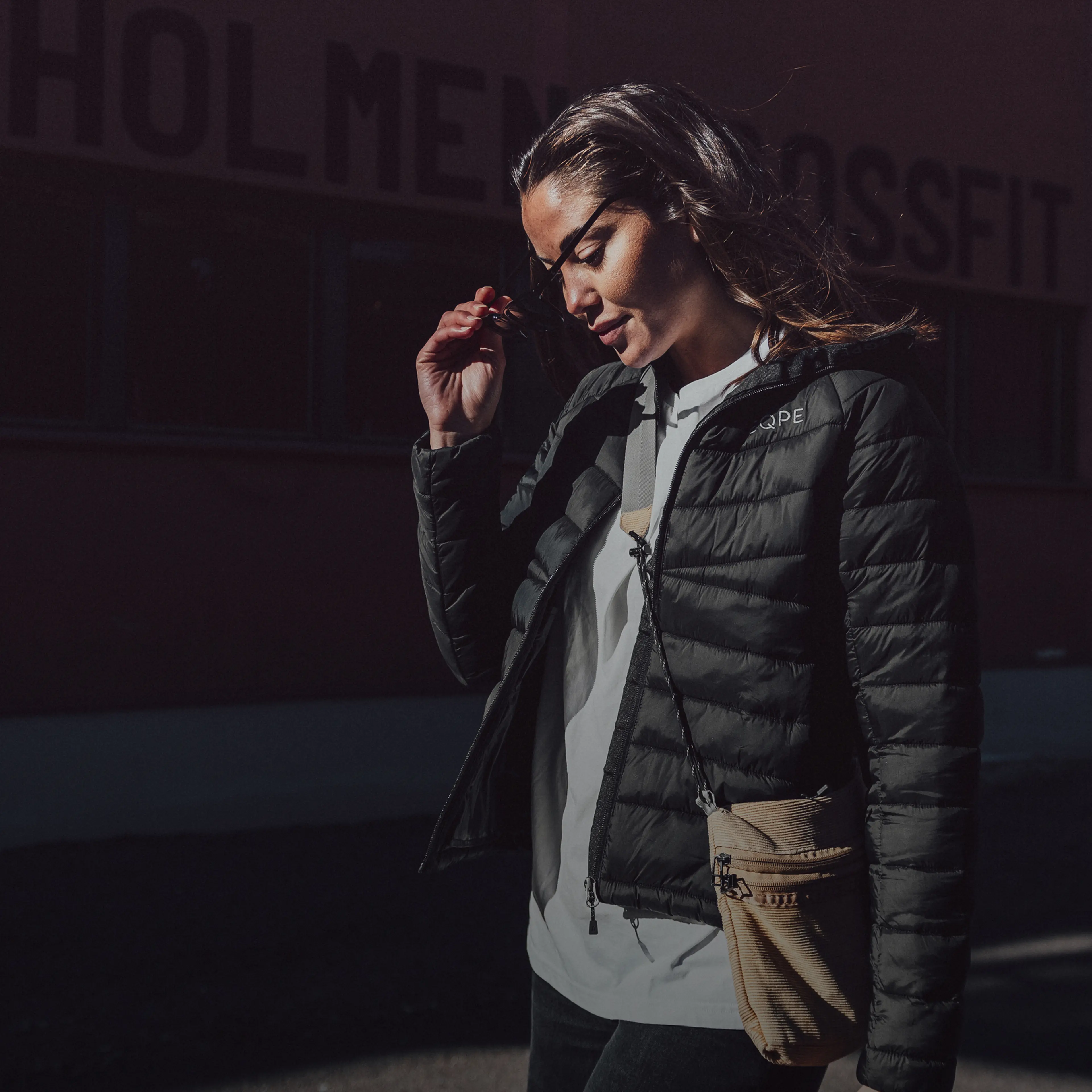 Woman in a black jacket and sunglasses outside, standing against a dark background with "HOLMEN CROSSFIT" partially visible.