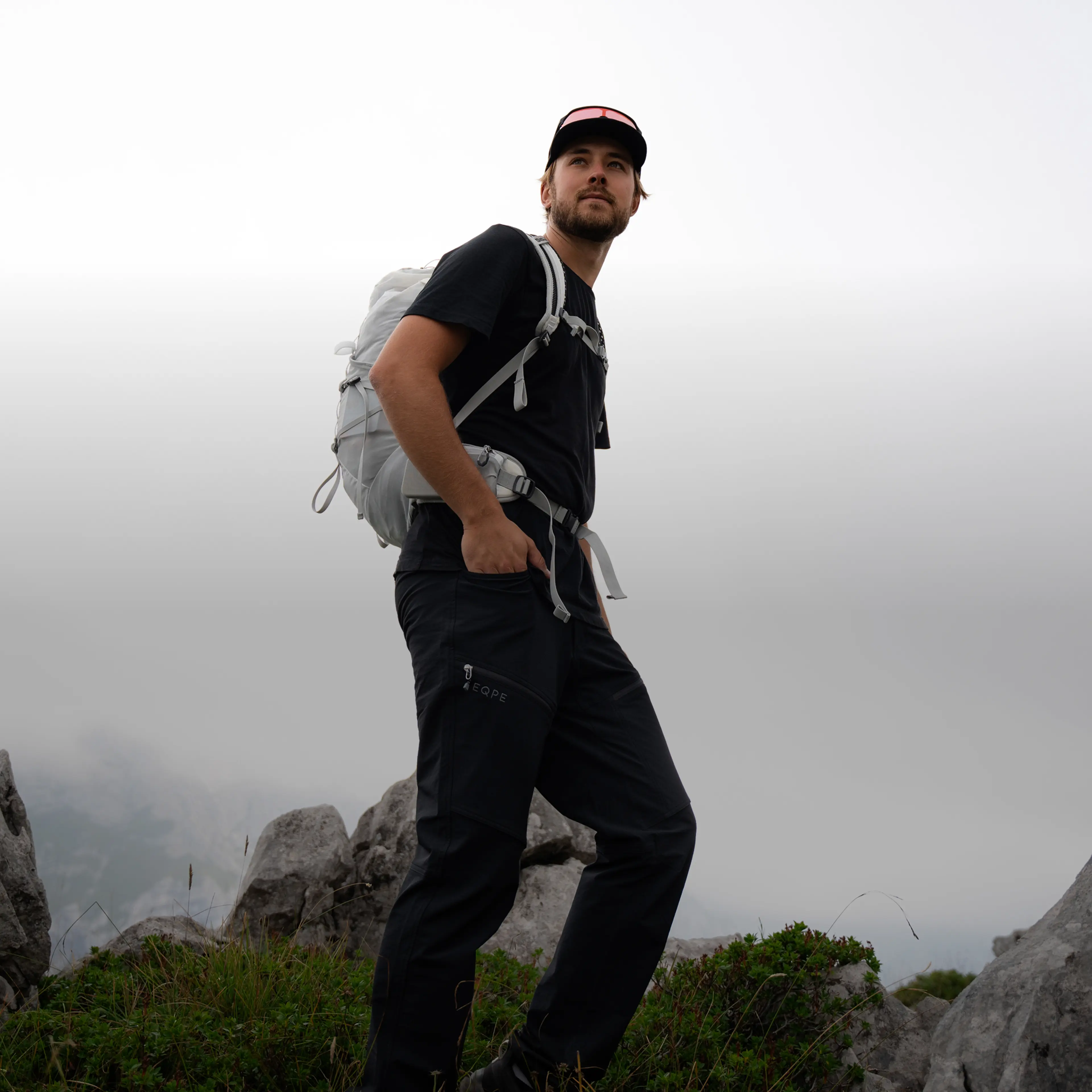 Man wearing a cap and backpack stands on a rocky, grassy hill with a cloudy sky in the background.