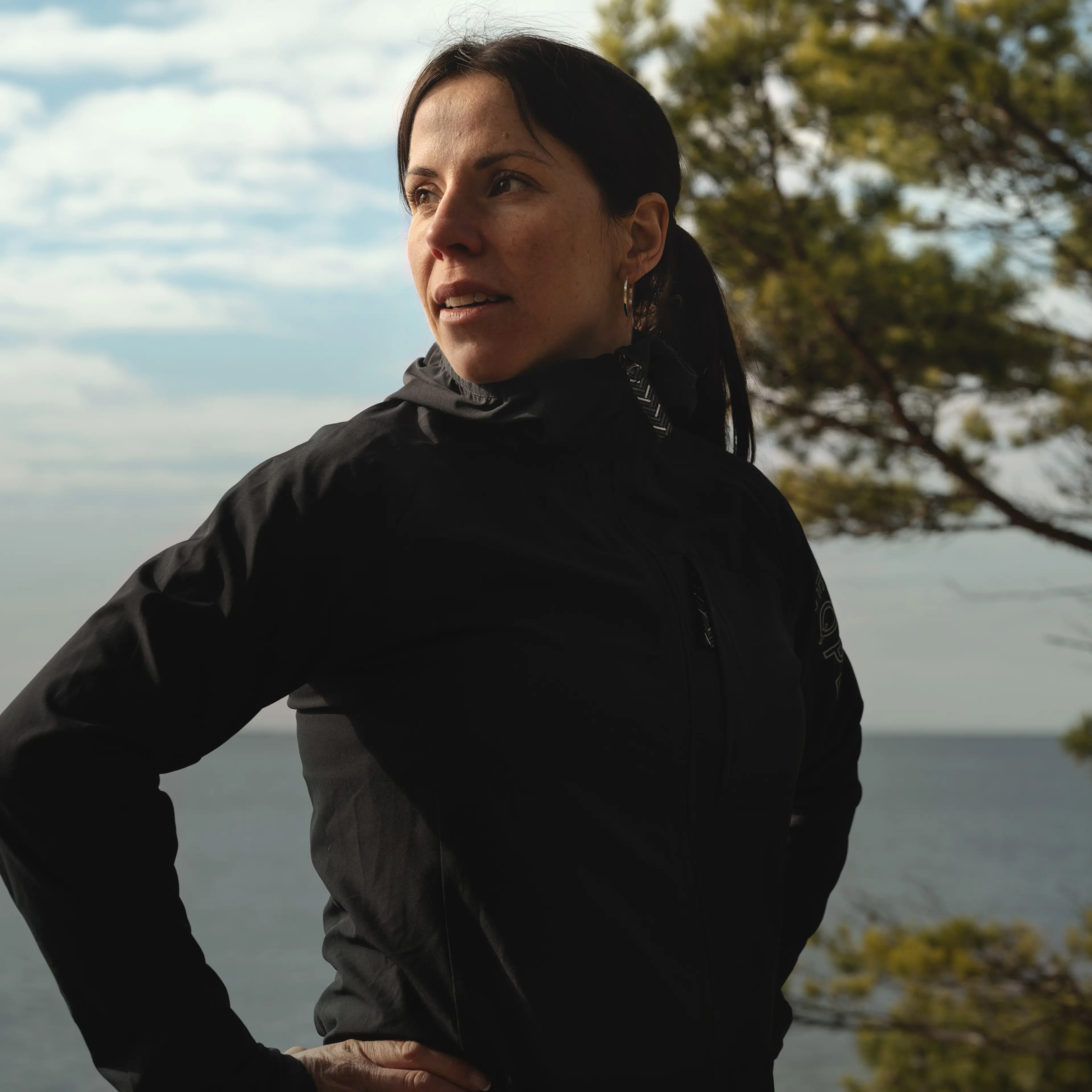 A woman in a black jacket stands outdoors by the sea, looking to the side with trees in the background under a cloudy sky.