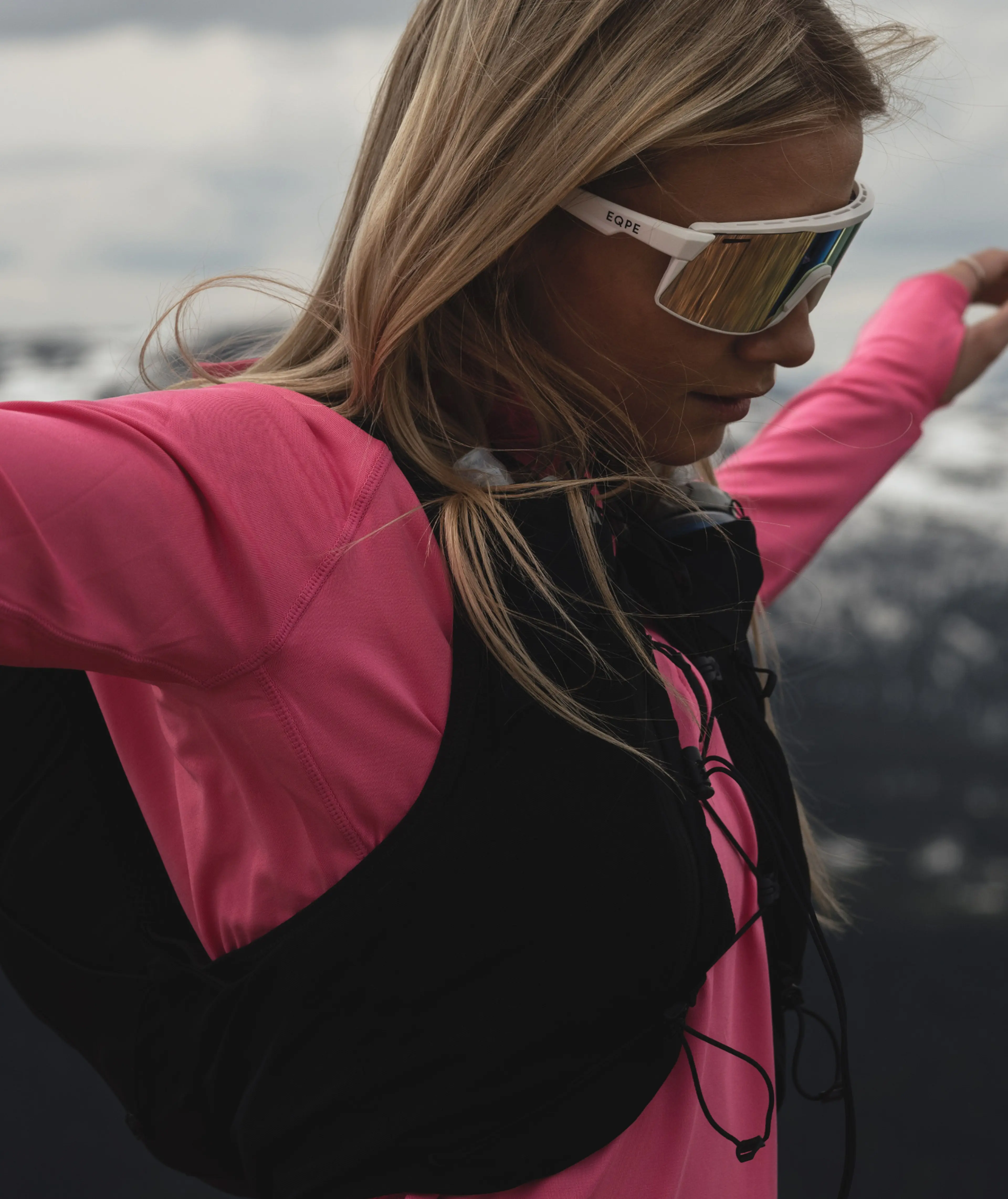Woman in a pink running shirt and sunglasses, standing outdoors with a mountainous background.