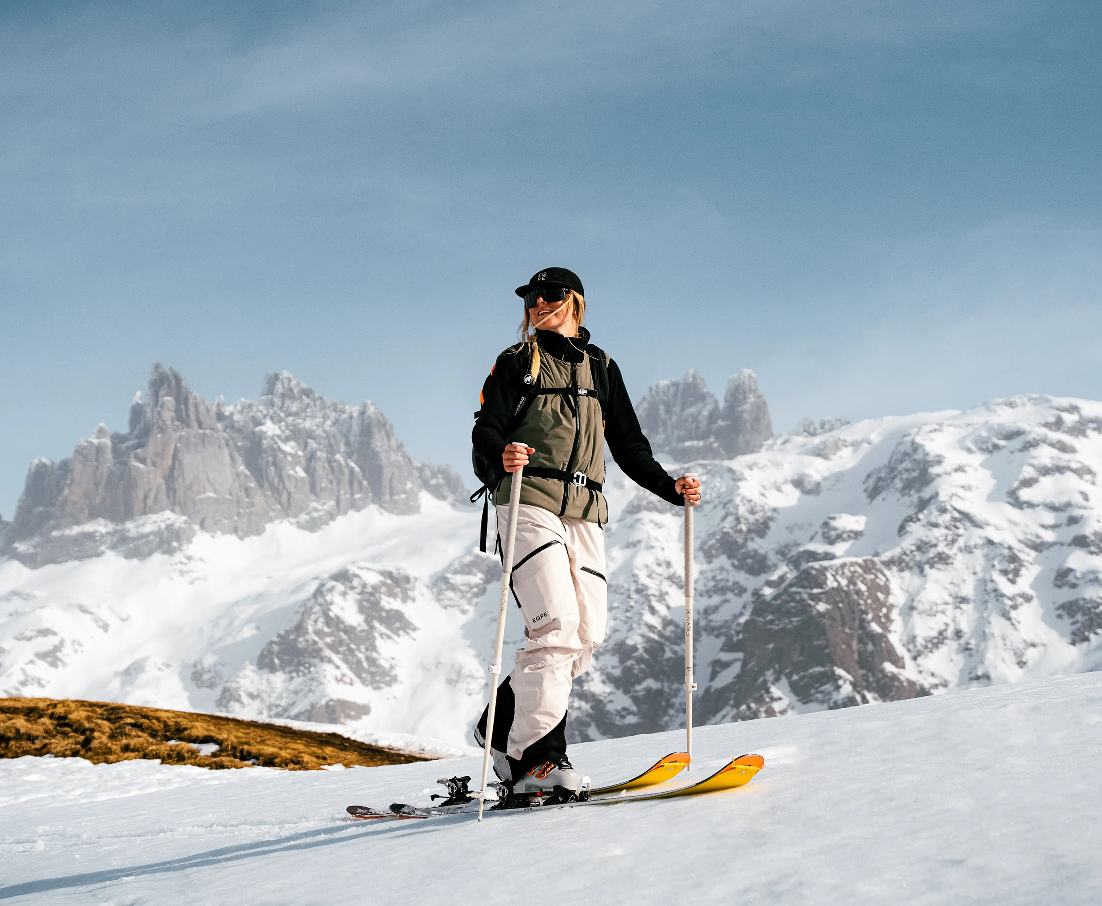 A skier in winter gear stands on a snowy slope with ski poles, set against a stunning backdrop of snow-capped mountains and a clear blue sky.