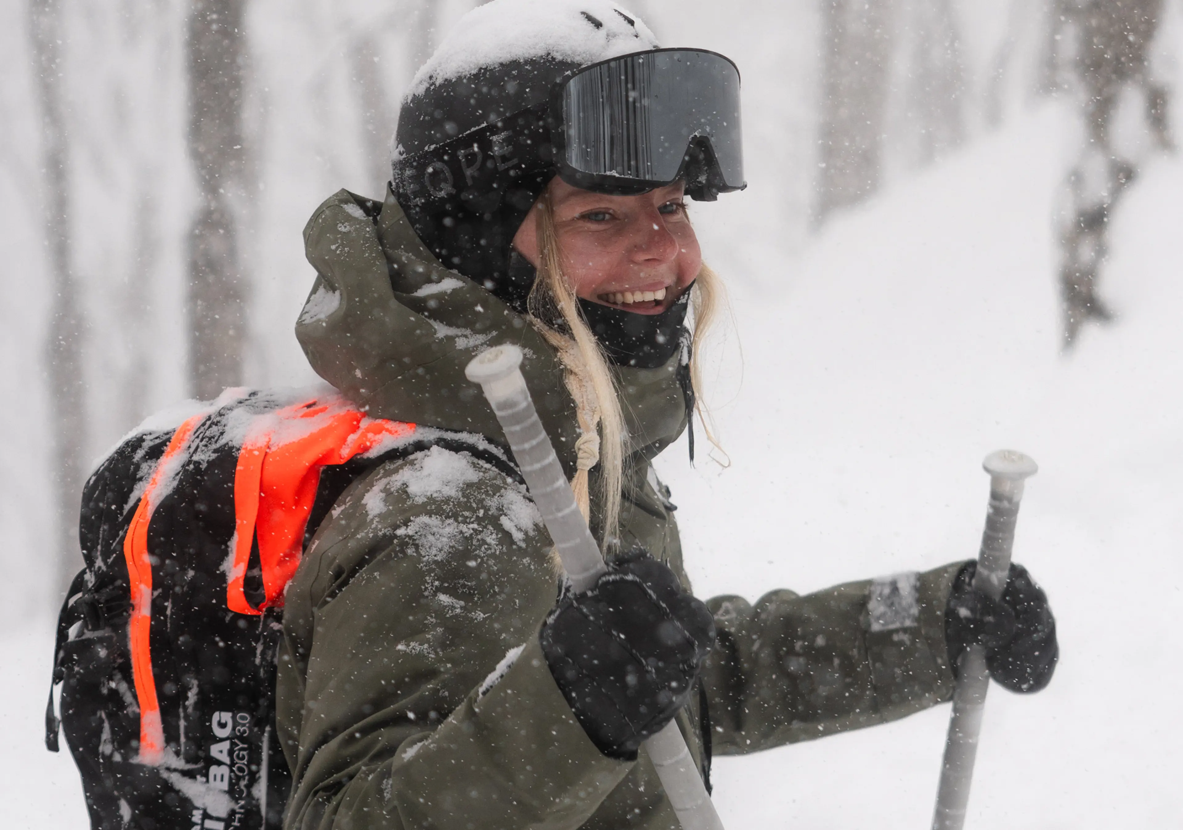 A smiling skier in snowy weather, wearing a helmet and goggles, holding ski poles, with a bright orange-accented backpack.