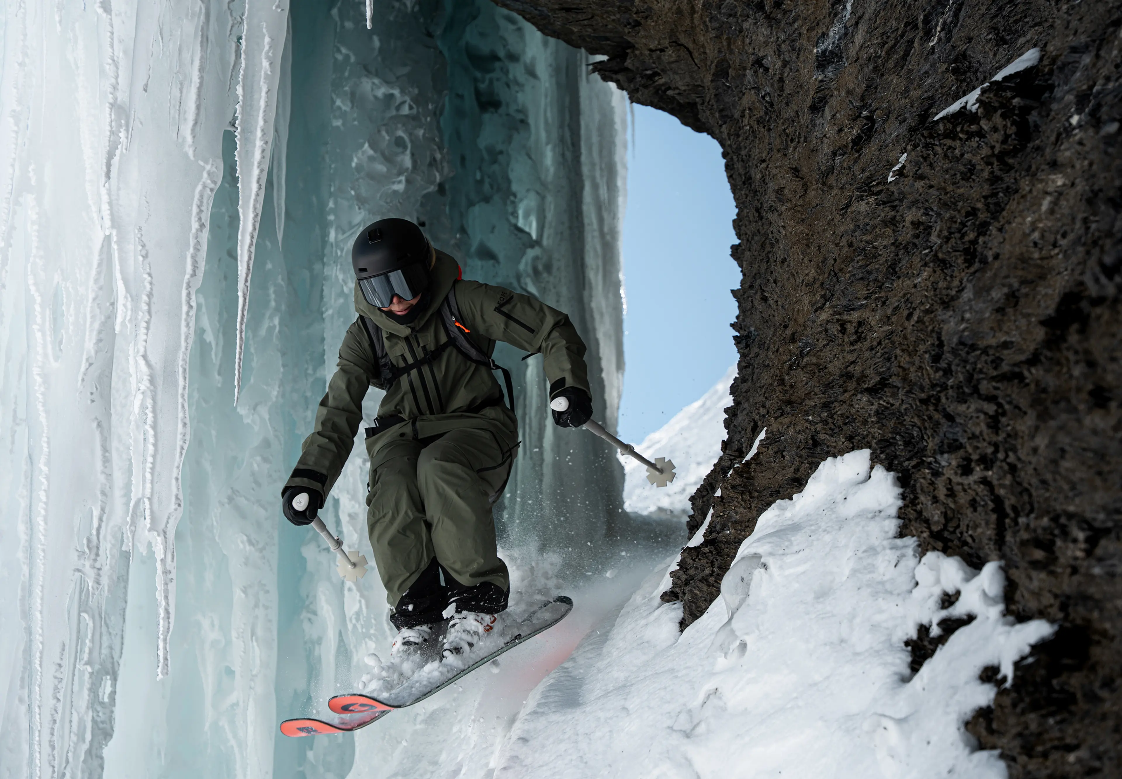 Skier in dark gear skiing down a snowy, narrow mountain passage with icy walls and a clear blue sky in the background.