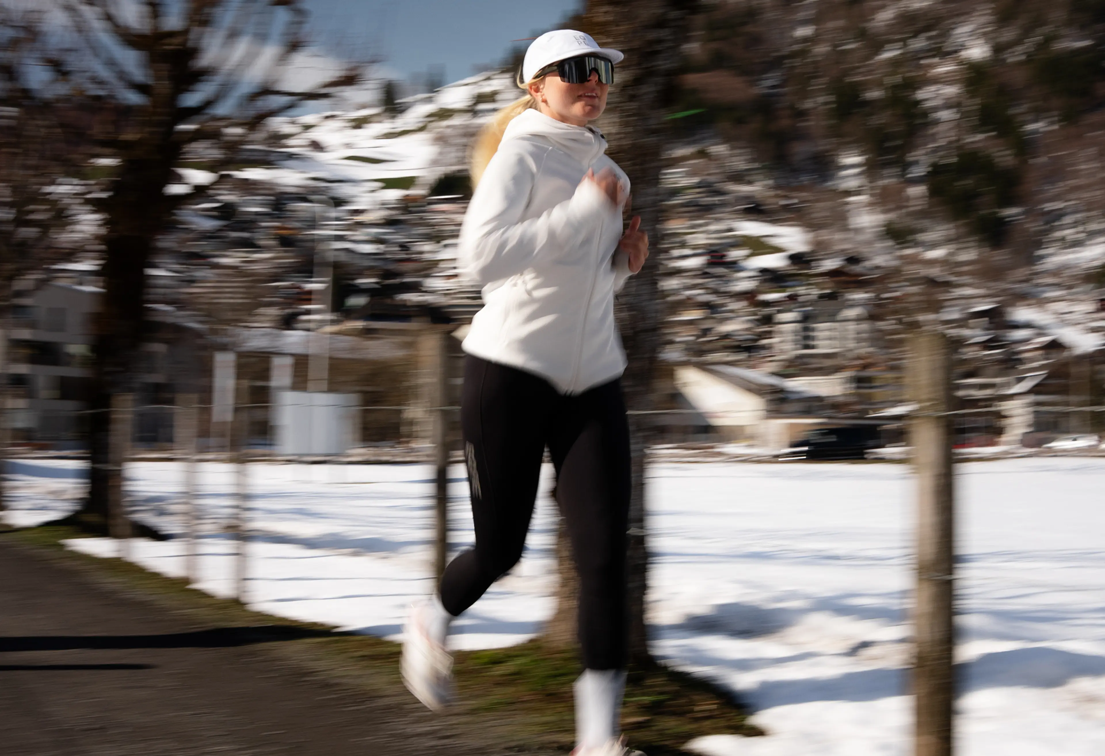Person jogging on a snowy path, dressed in white, with blurred trees and buildings in the background.