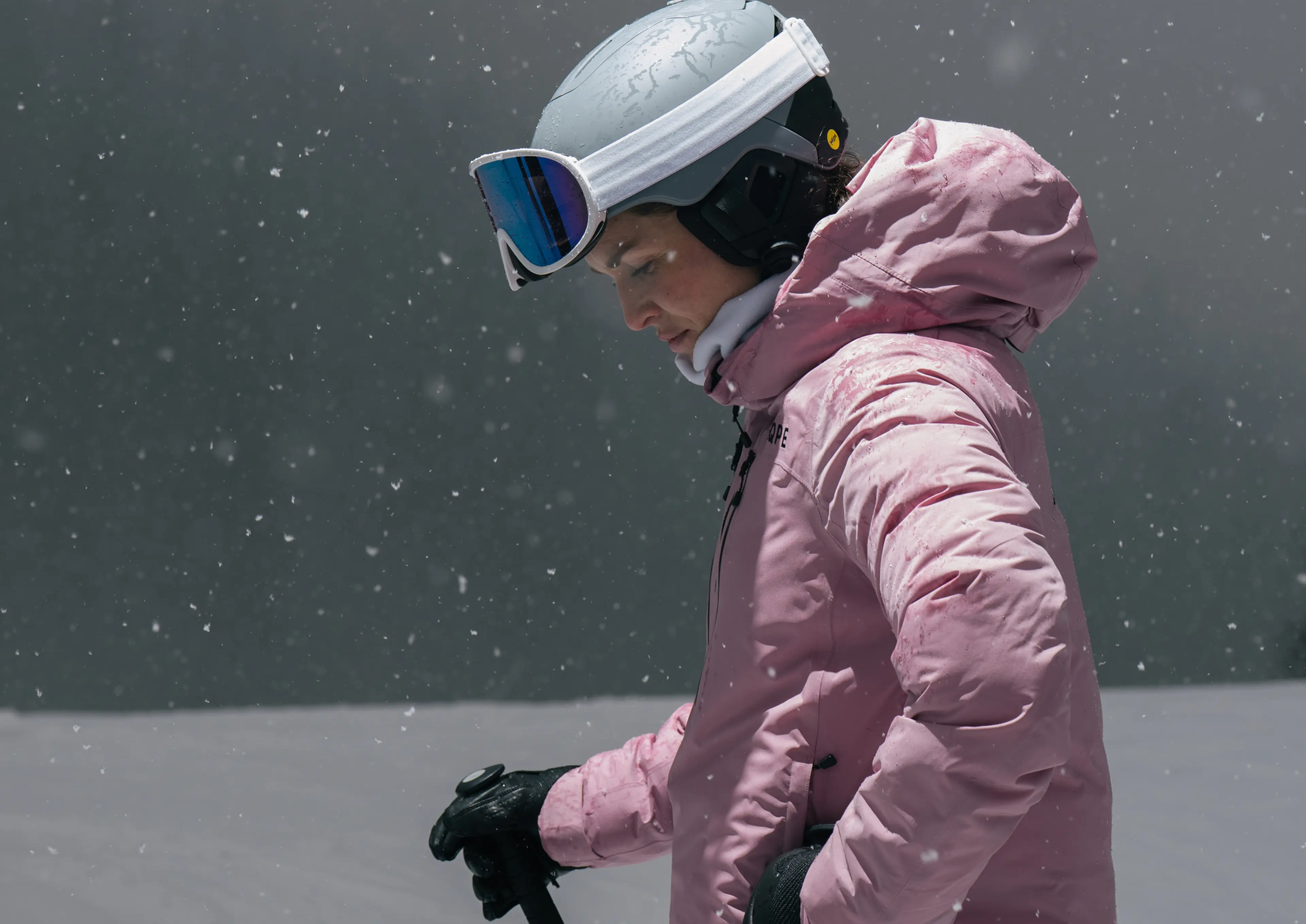 A skier in a pink jacket and helmet stands in the snow, looking down. Snowflakes gently fall against a grey background.