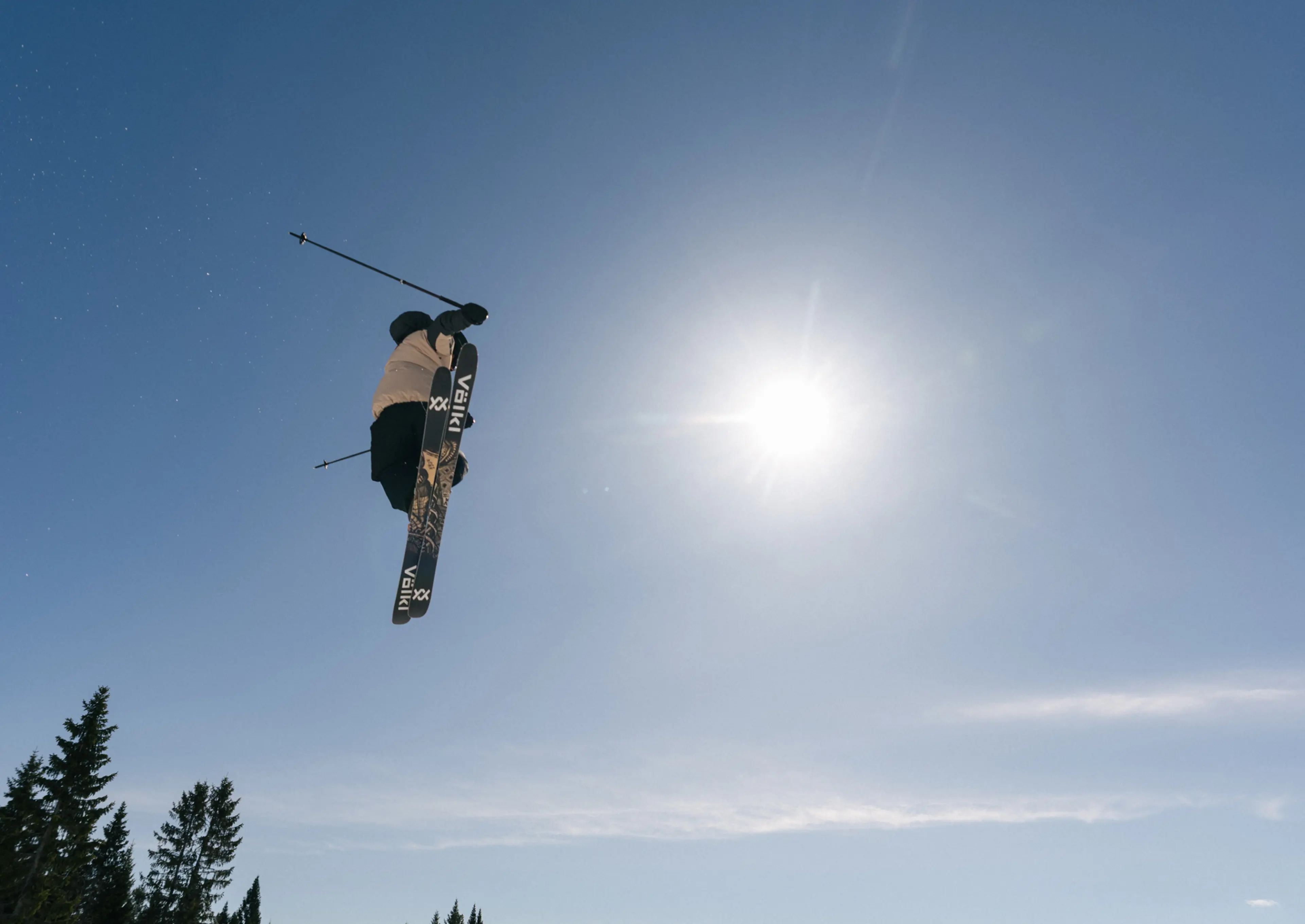 A skier performs a mid-air trick under a clear blue sky, with bright sun and trees below.