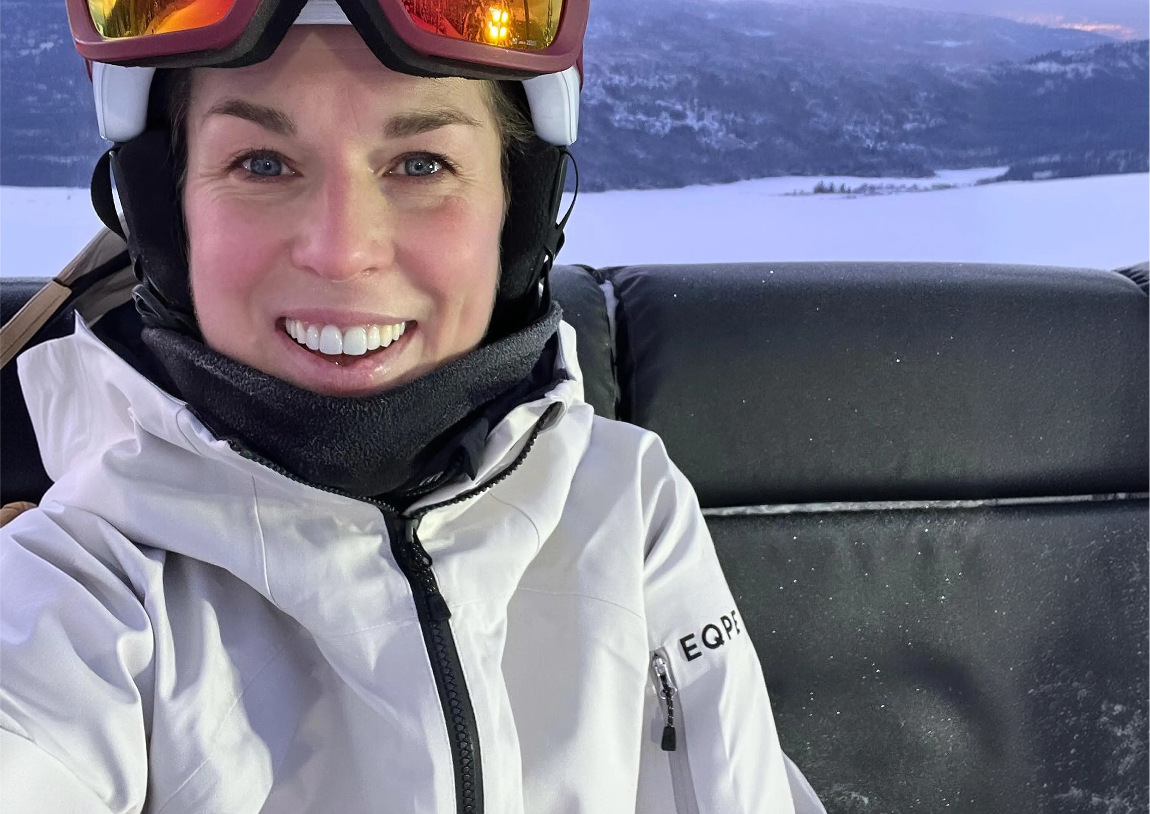 Smiling person in ski gear with goggles, sitting on a ski lift with a snowy mountain landscape in the background.