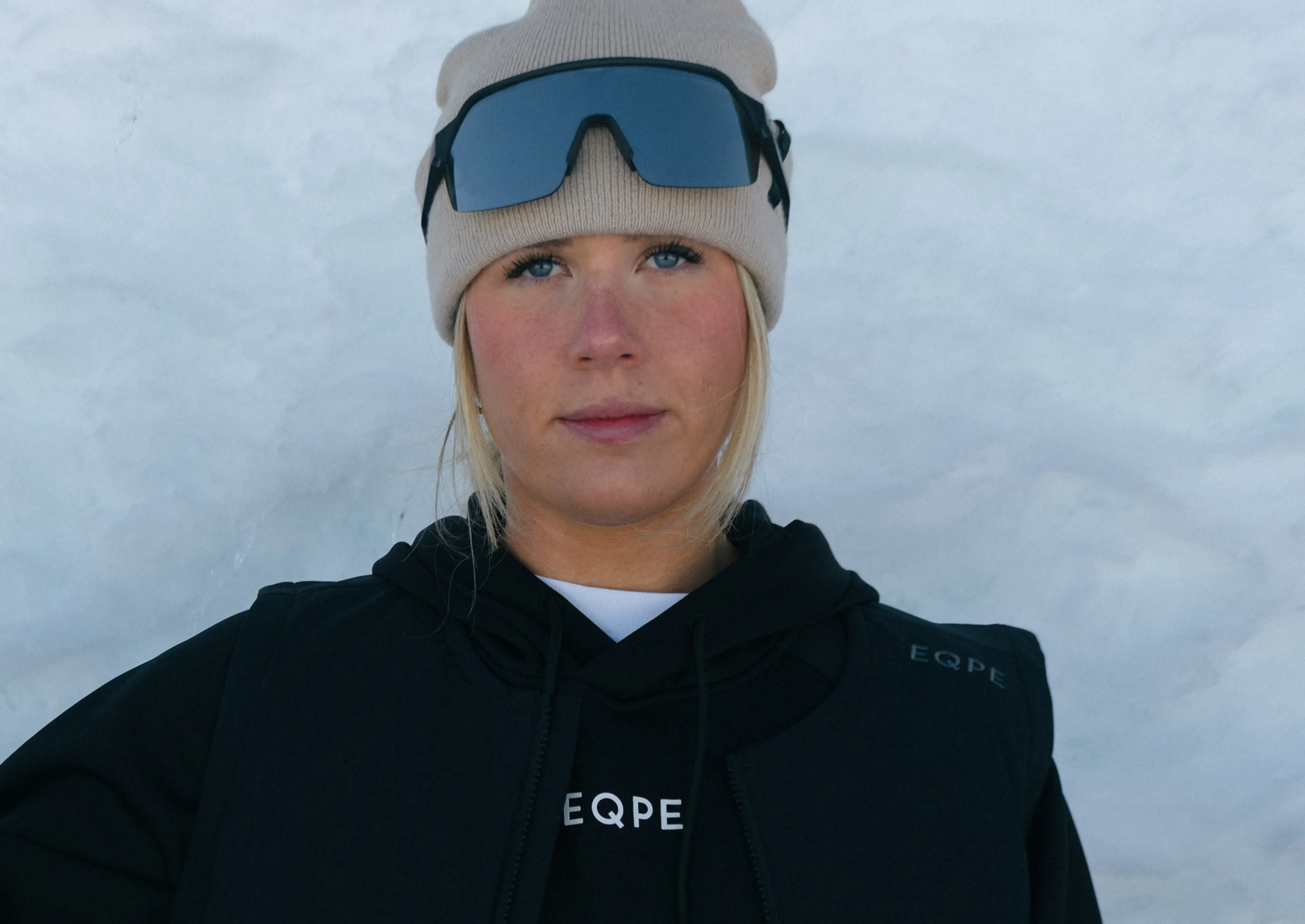 Person wearing a beige beanie, black tinted goggles, and a black EQPE jacket in front of a snowy background.
