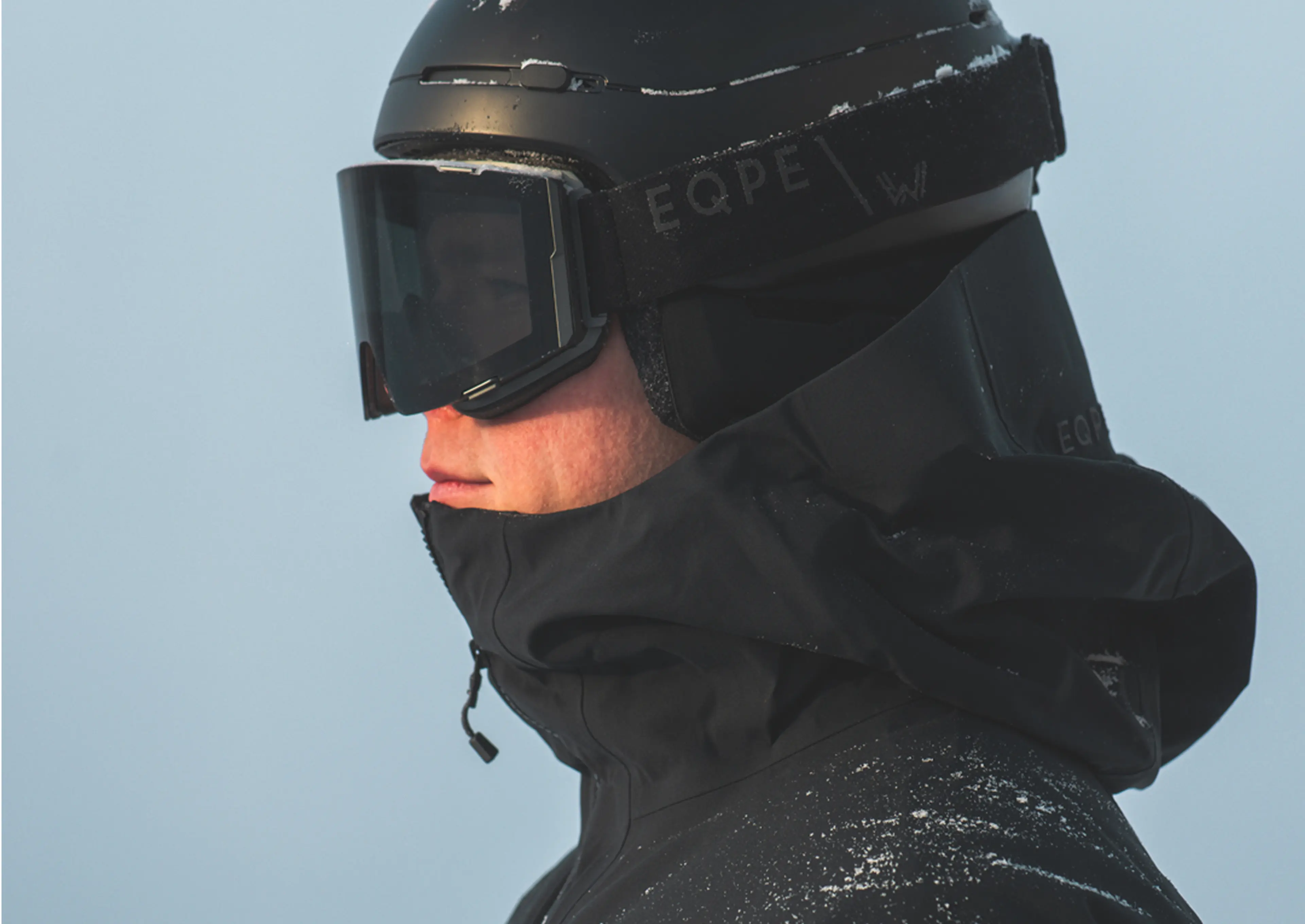 Person in a black helmet and ski goggles with snow on their jacket, standing against a snowy backdrop.