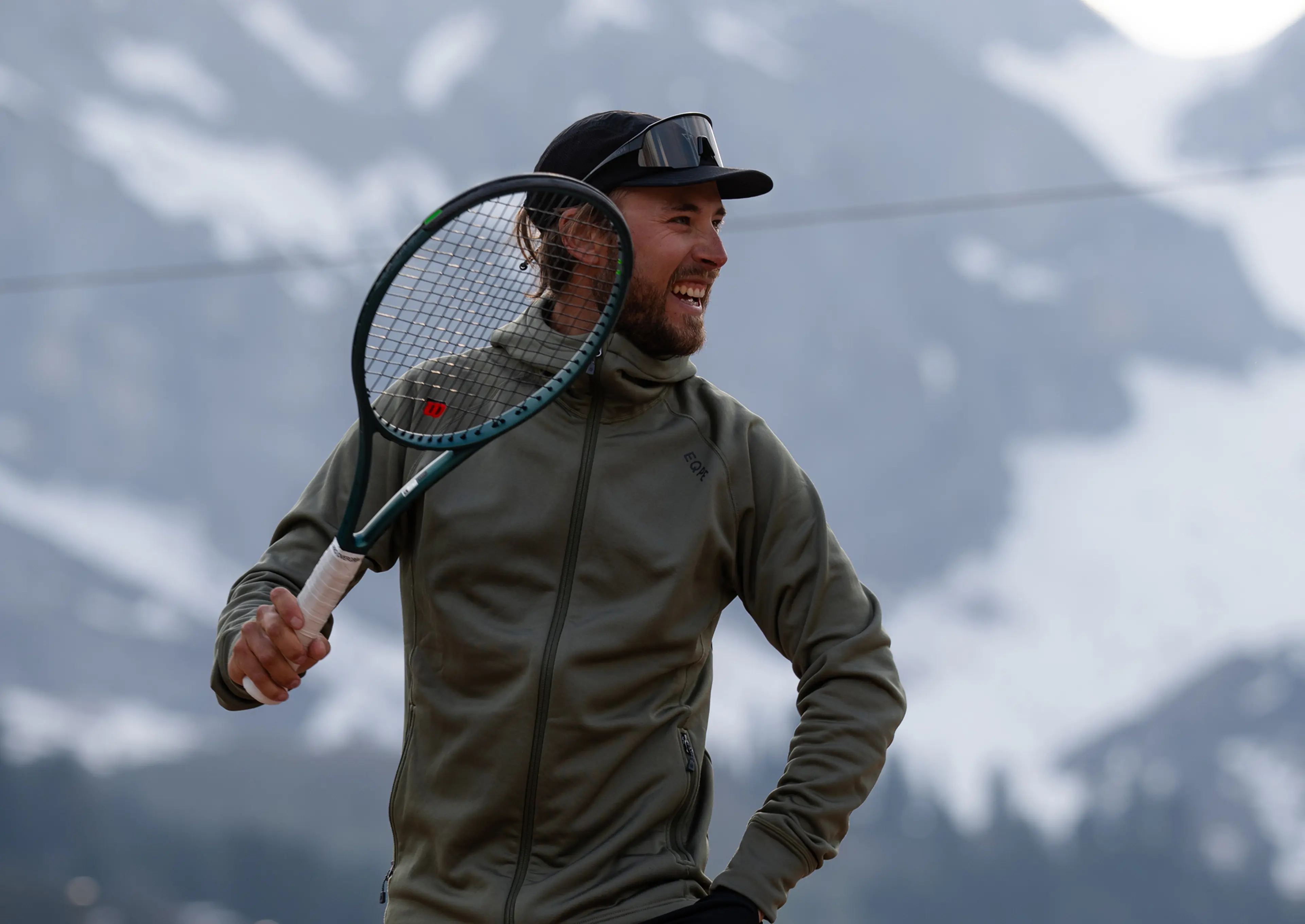A smiling man in a cap and jacket playfully holds a tennis racket, with snowy mountains in the background.