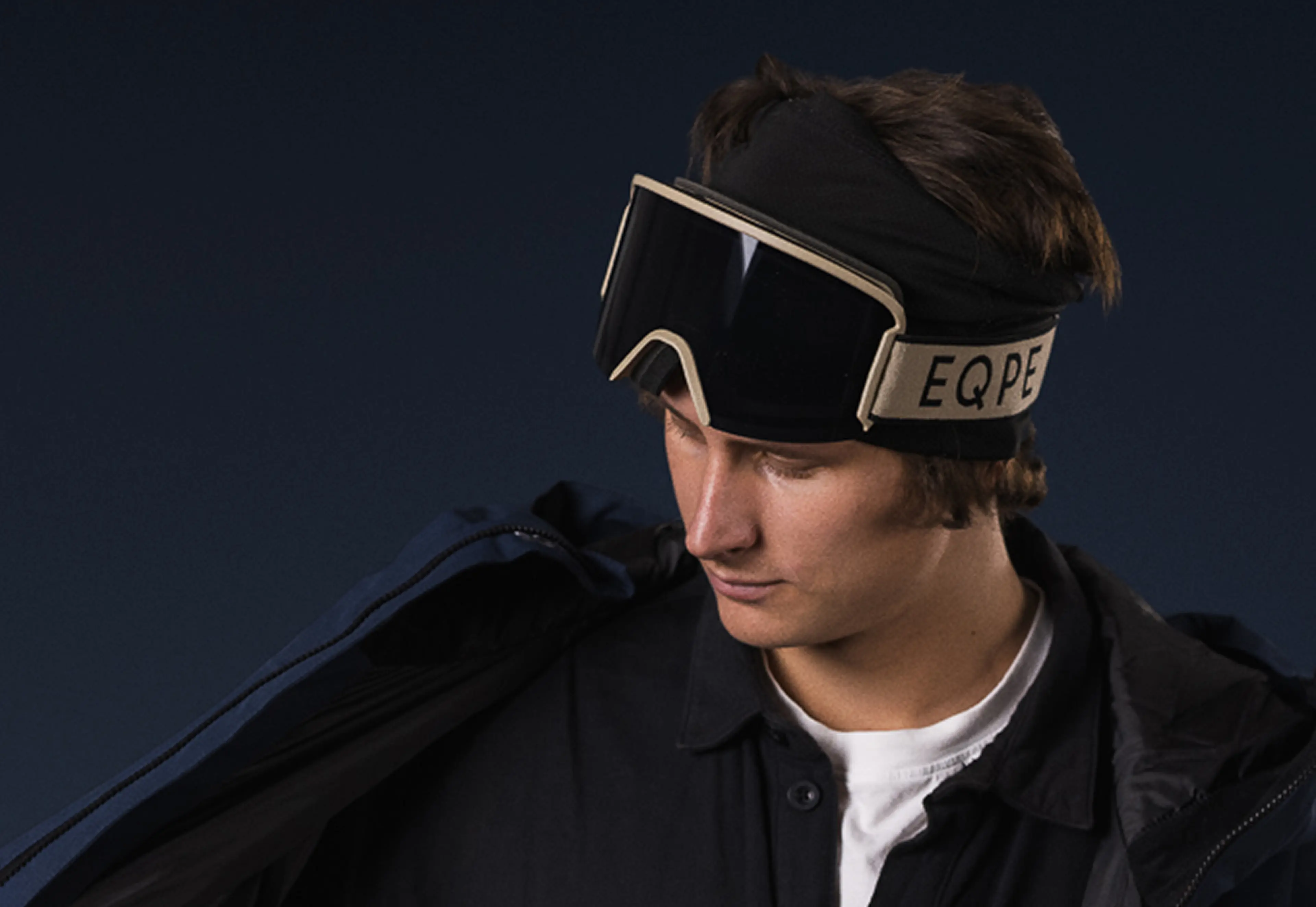 Person wearing ski goggles and black jacket, looking down against a dark background.
