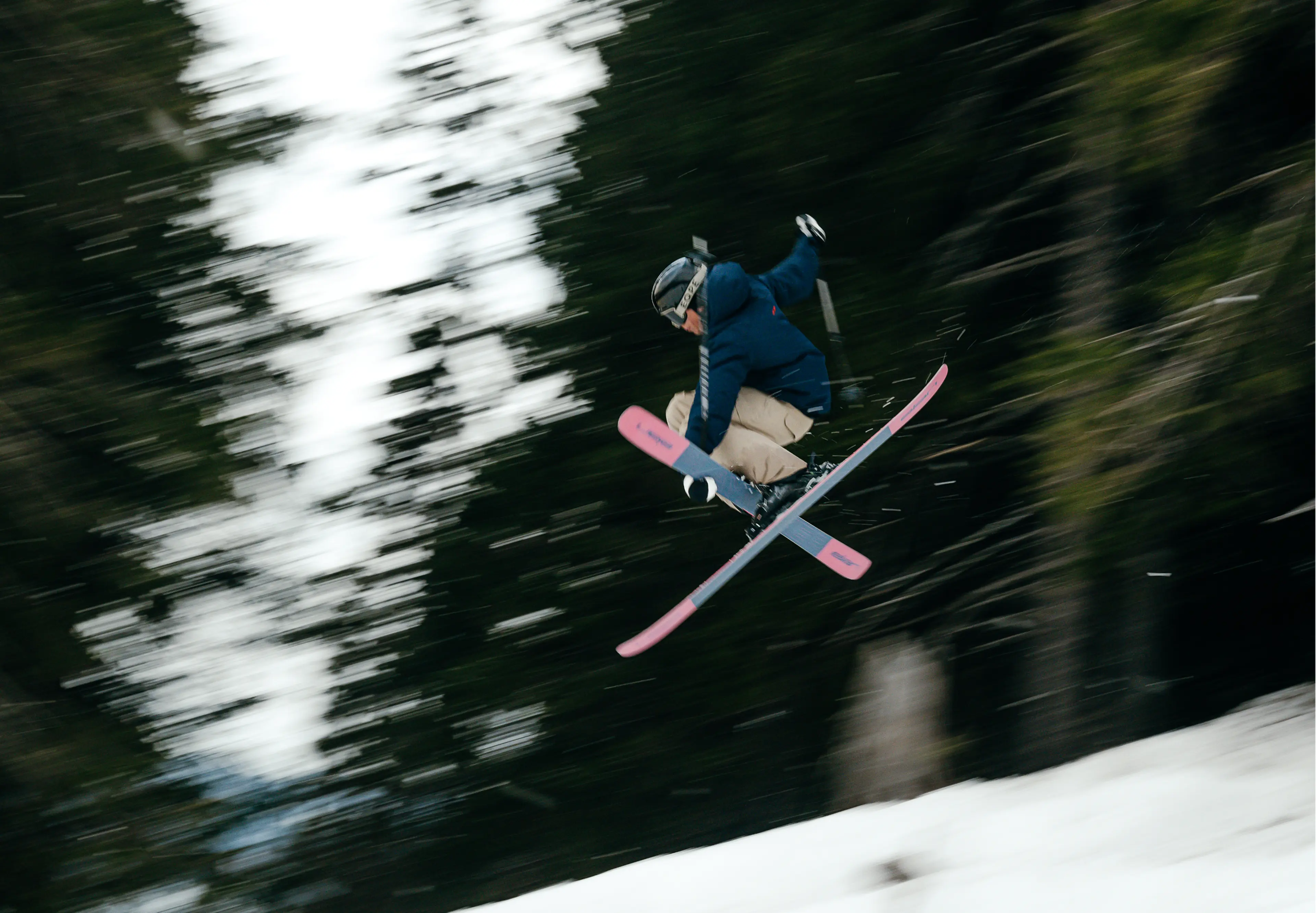Skier in mid-air with pink skis, wearing a helmet, blue jacket, and beige pants, performing a trick against a blurred forest background.