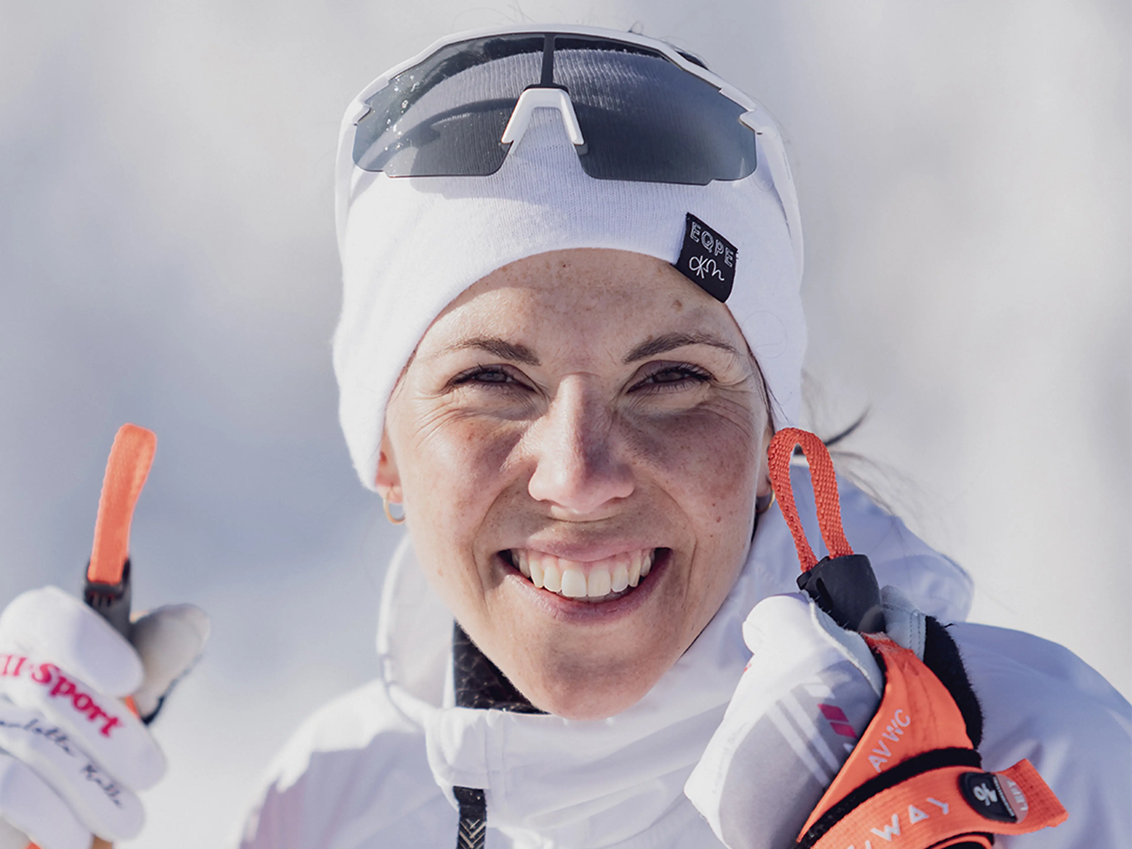 Smiling person in winter gear with white hat and sunglasses, holding ski poles, against a snowy background.